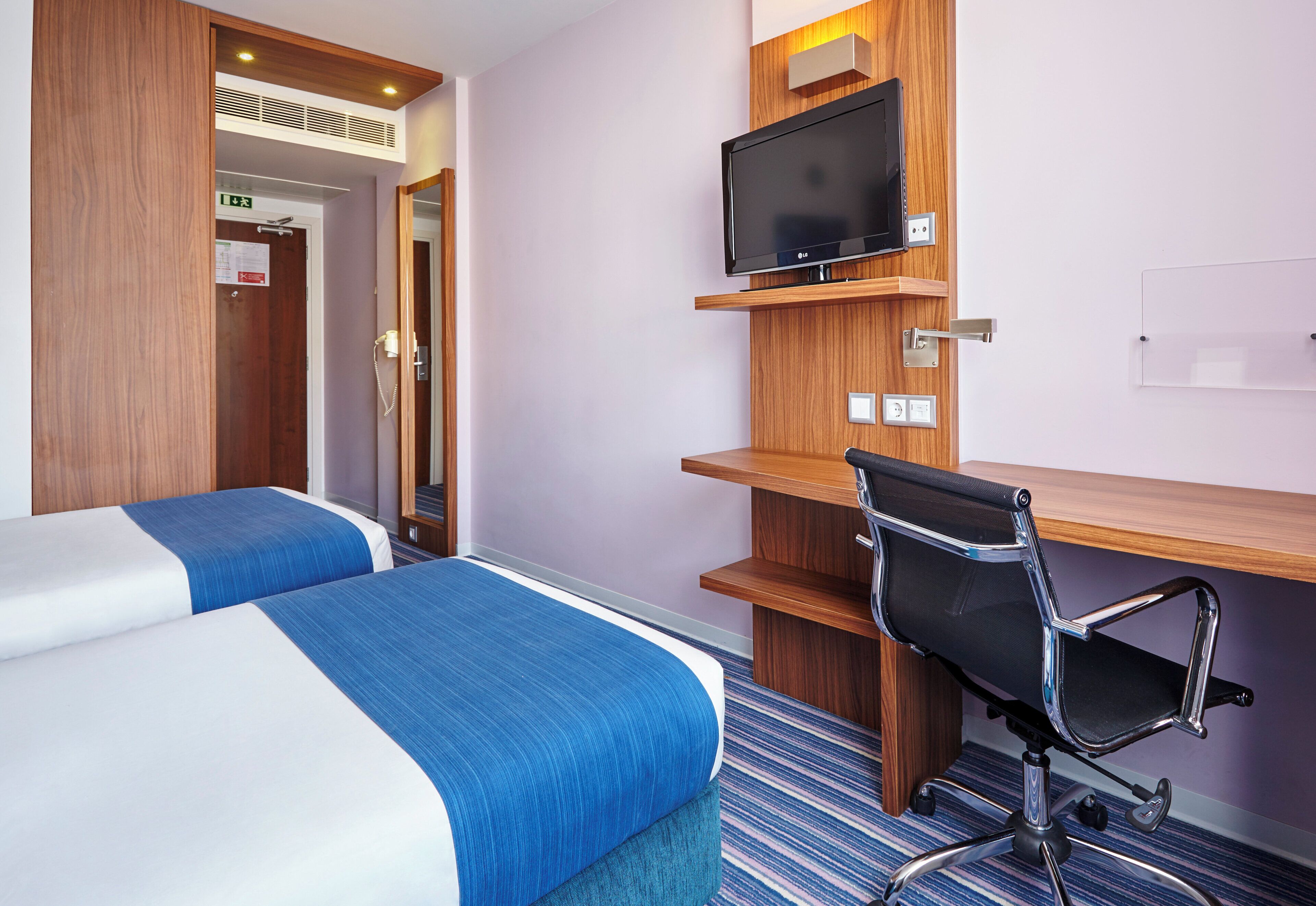 Ein Hotelzimmer mit zwei Betten, einem Schreibtisch, einem Computerbildschirm, einem Stuhl und einem Telefon.