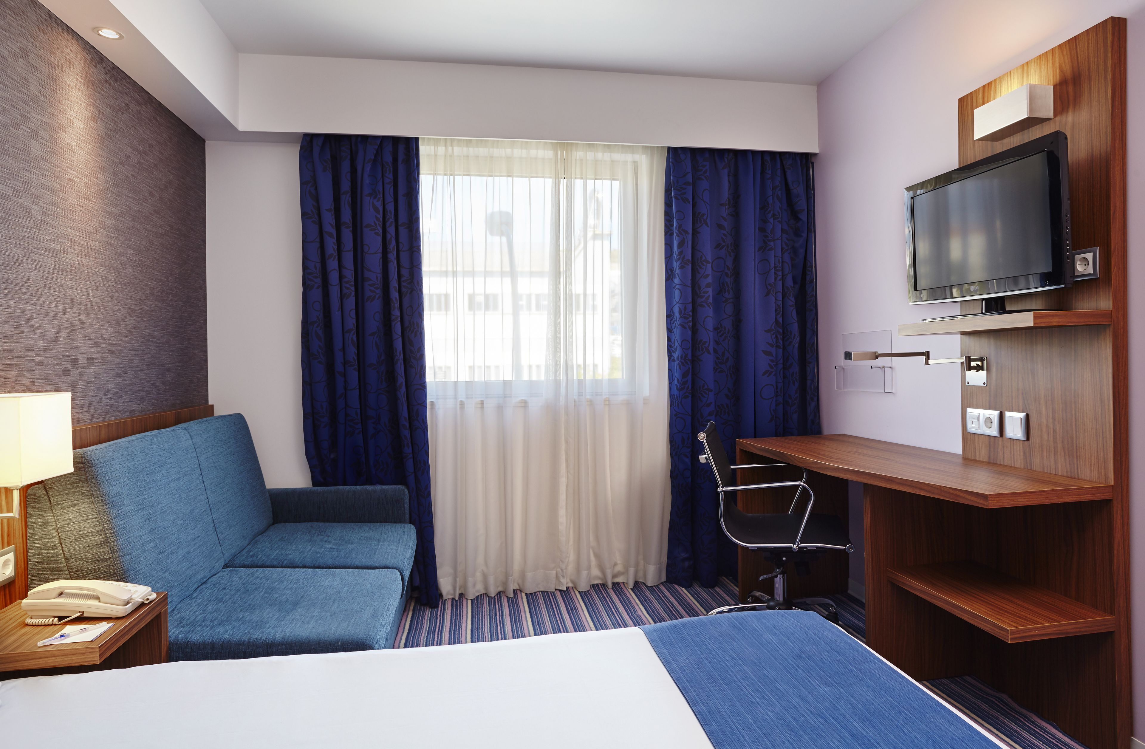 A hotel room with a bed, a desk, a chair, a TV, and a window with curtains.