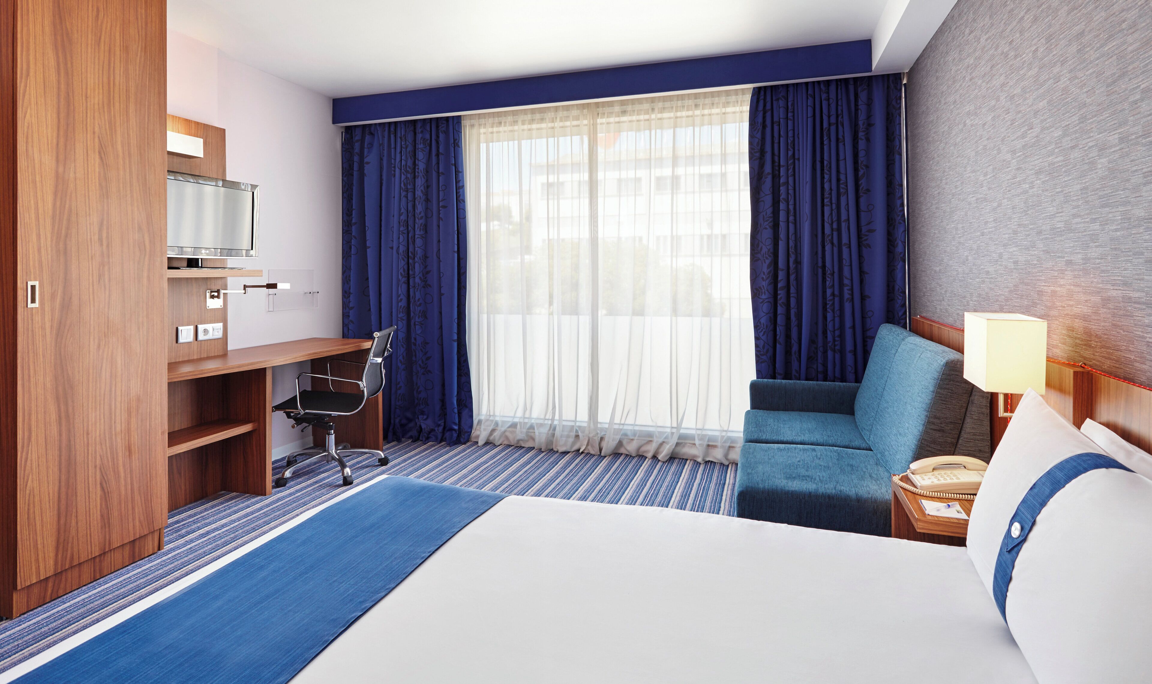 A hotel room with a bed, a desk, a chair, a TV, and a window with curtains.