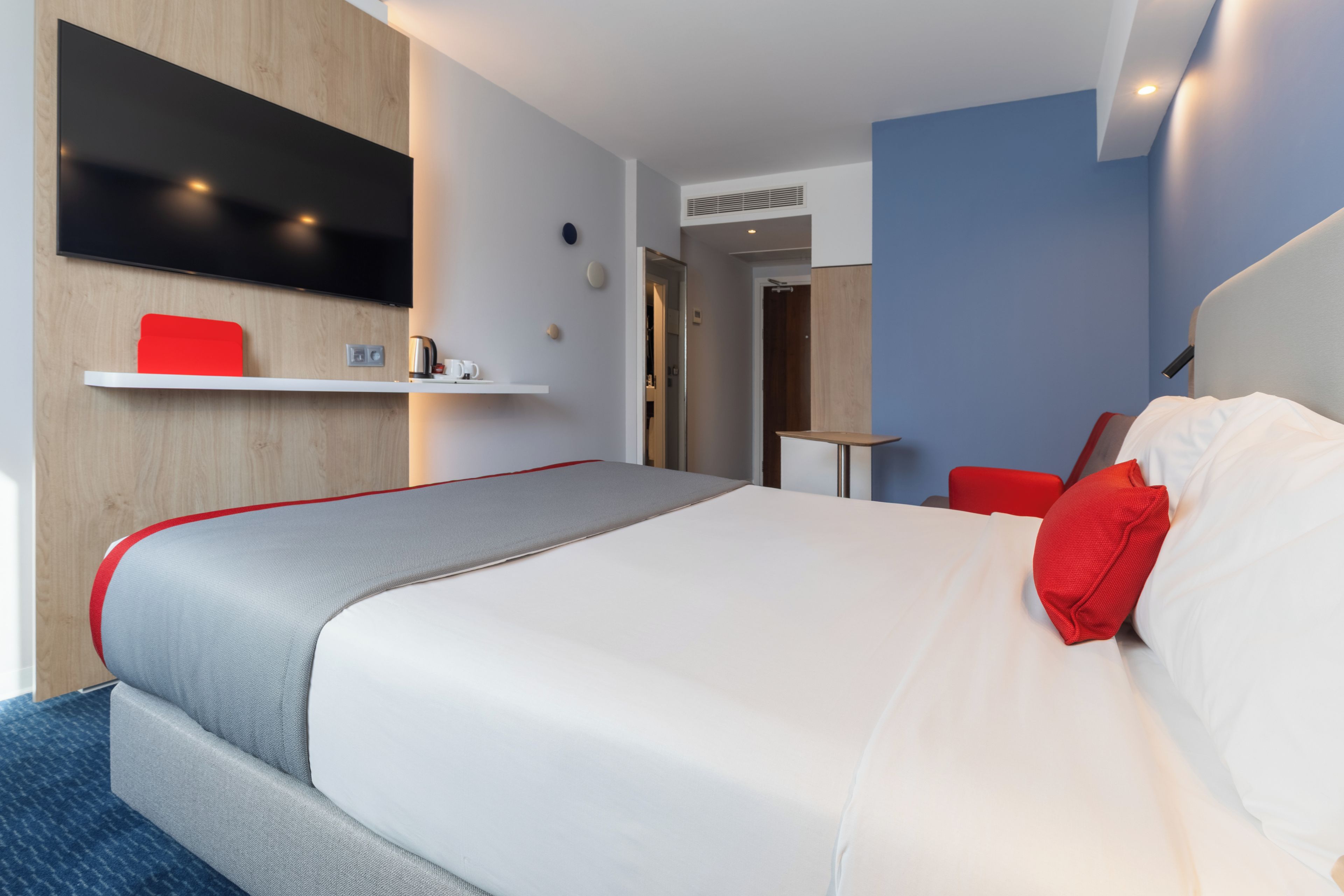 Quarto de hotel moderno com uma cama grande, uma televisão de tela plana na parede, uma pequena mesa e uma cadeira vermelha.