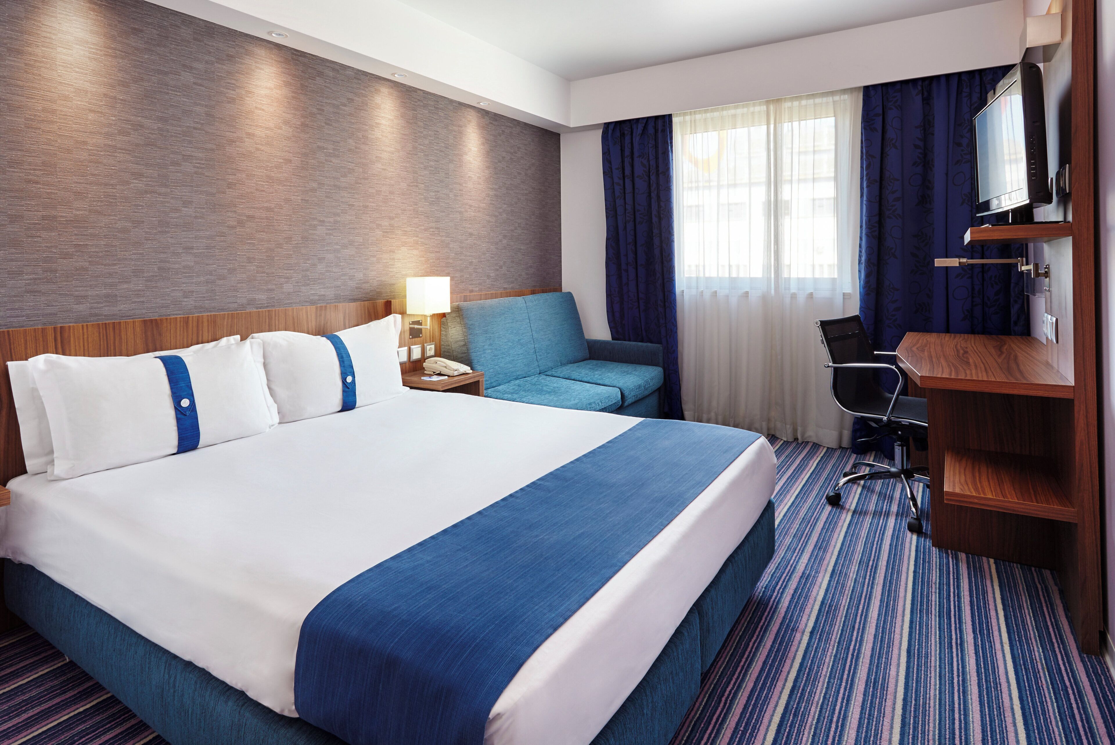 A hotel room with a large bed, a desk, a chair, a TV, and a window with curtains.