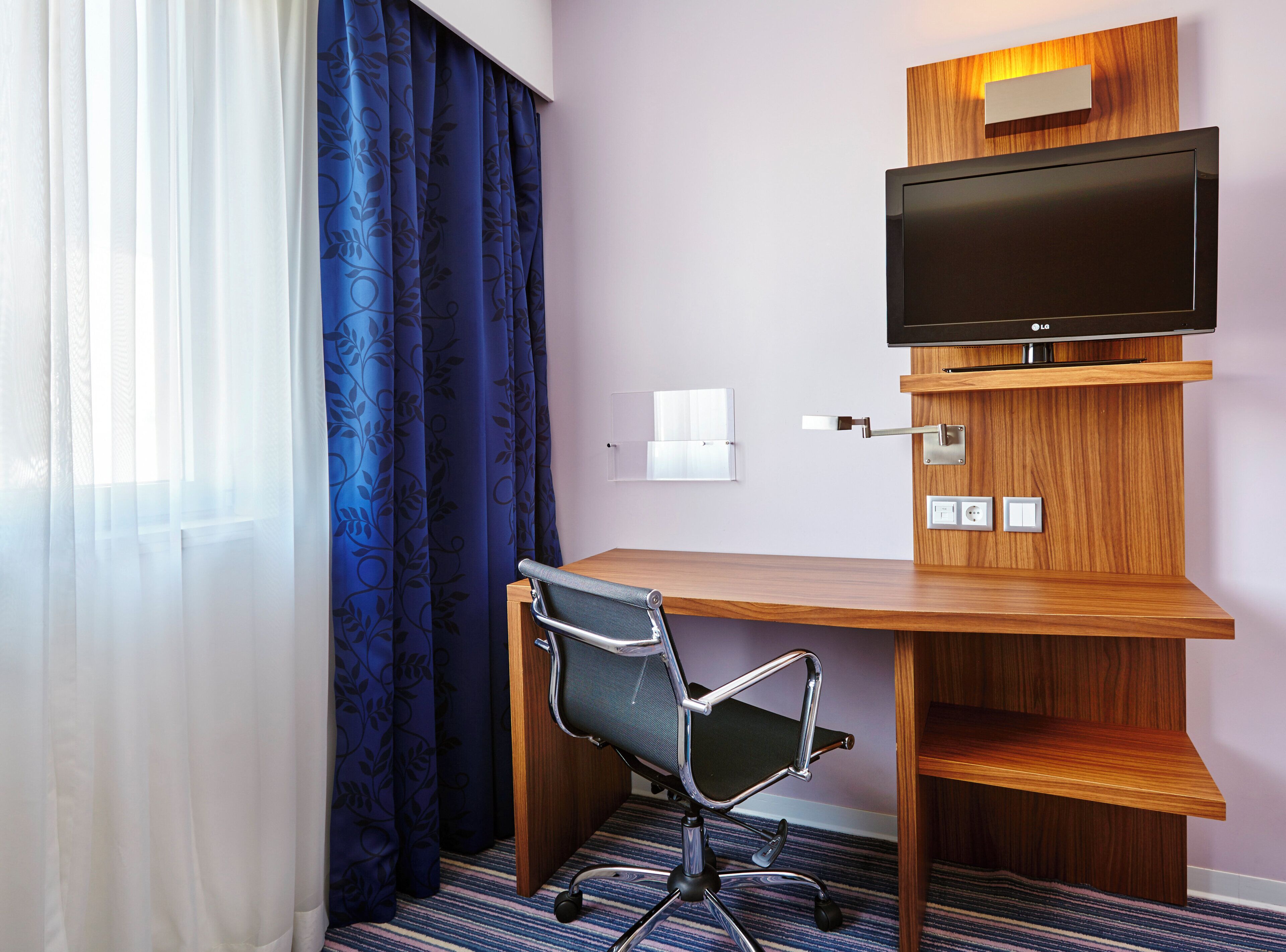 Habitación de hotel con un escritorio de madera, una silla, un televisor montado en un panel de madera y una ventana con cortinas azules.
