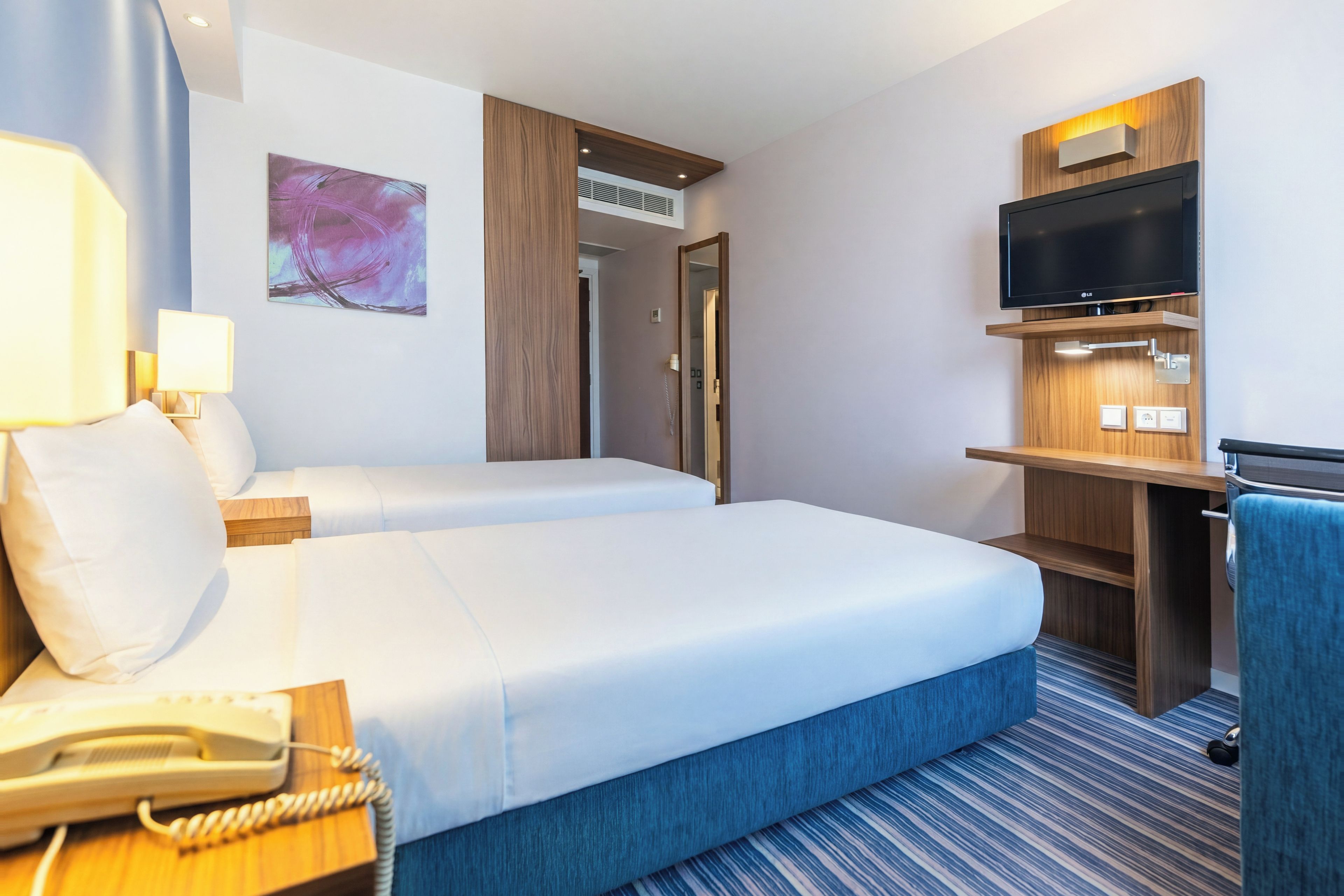 A hotel room with two beds, a desk with a telephone, a flat-screen TV, and a painting on the wall.