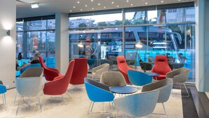 A modern indoor seating area with a mix of red and blue chairs, a glass partition, and a view of a blue bus outside. - Holiday Inn Express Lisbon - Alfragide by IHG (Oeiras)