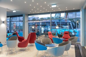 A modern indoor seating area with a mix of red and blue chairs, a glass partition, and a view of a blue bus outside. - Holiday Inn Express Lisbon - Alfragide by IHG (Oeiras)