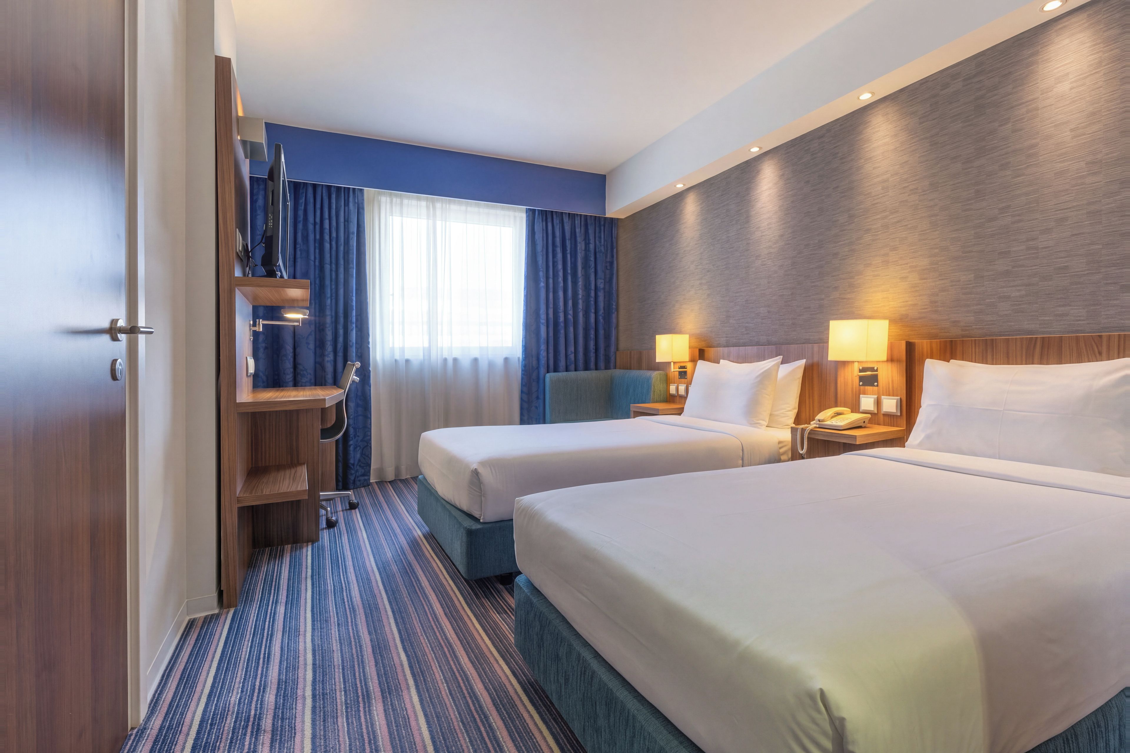 A hotel room with two beds, a desk, a chair, a TV, and a window with curtains.