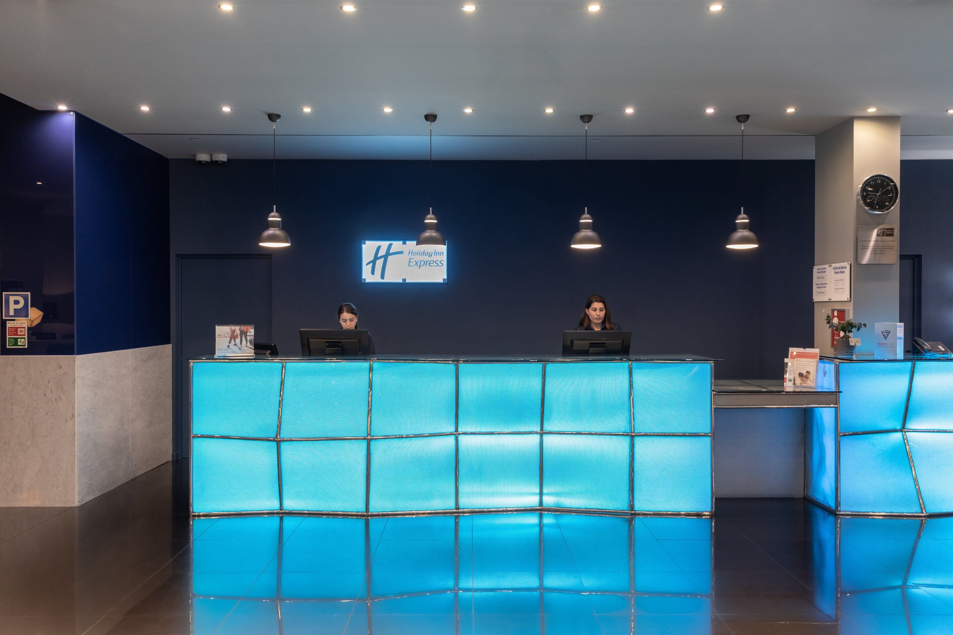 A Holiday Inn Express reception desk with a blue-tiled counter, overhead lighting, and a clock on the wall.