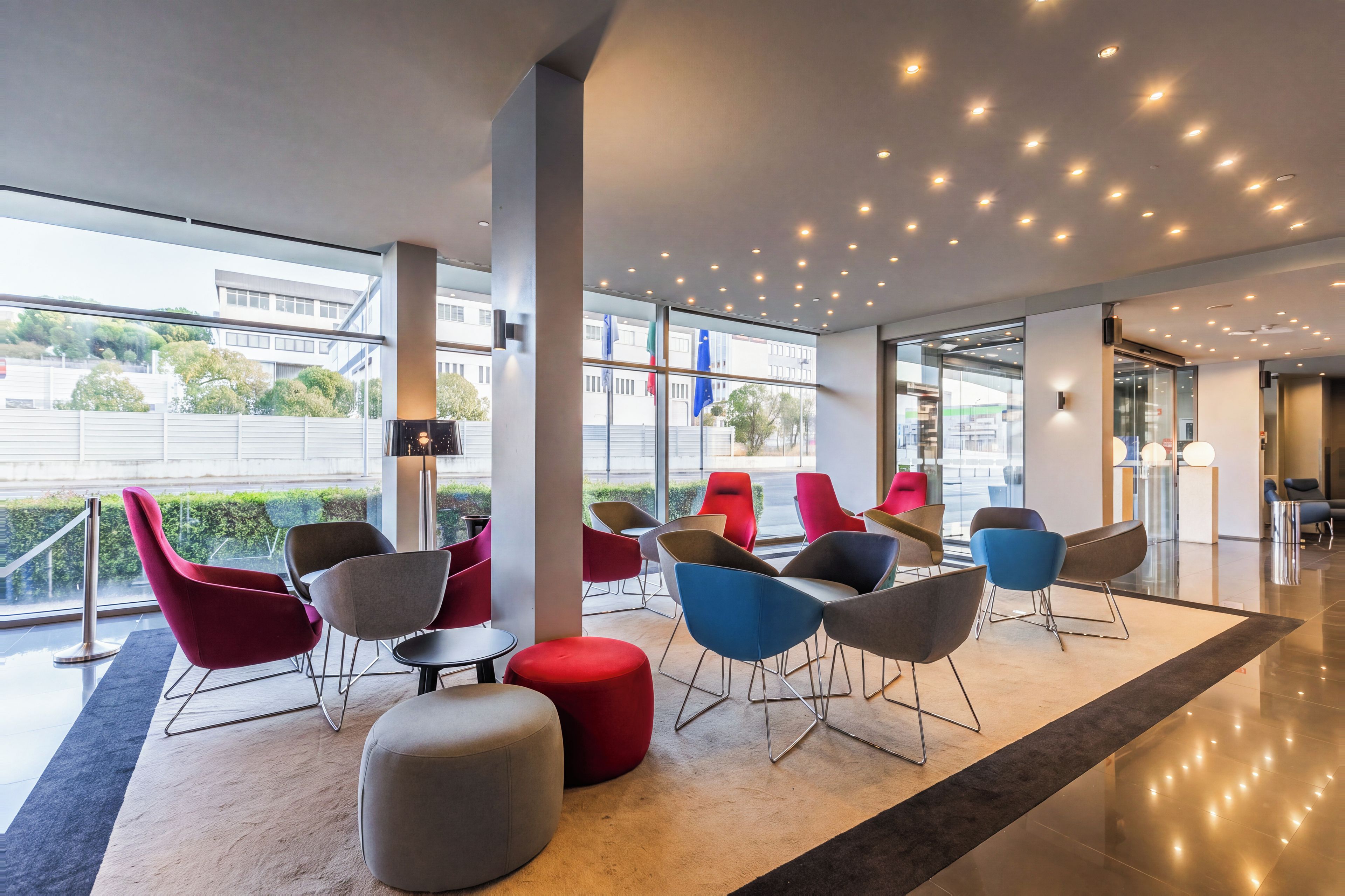 A modern indoor seating area with a variety of chairs and ottomans, large windows, and a ceiling with recessed lighting.