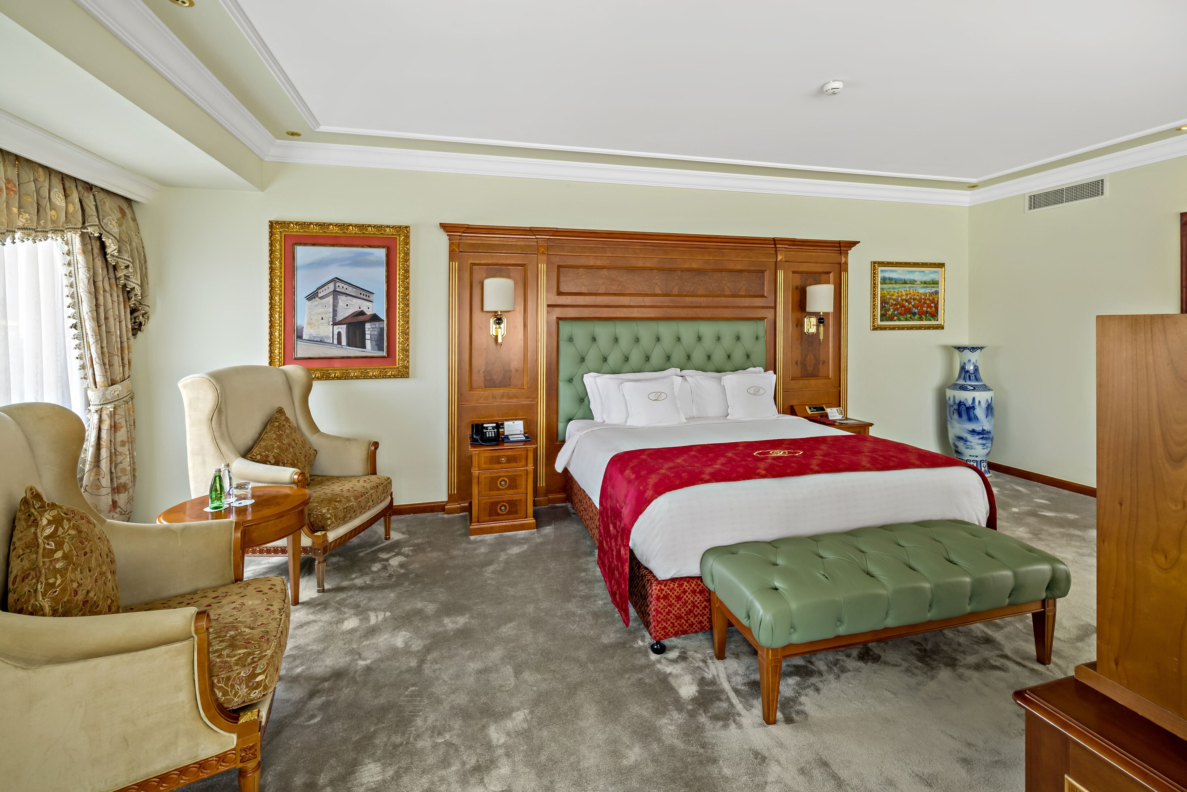 A hotel room with a large bed, a green upholstered bench, a wooden nightstand, a chair, and a large window with curtains.