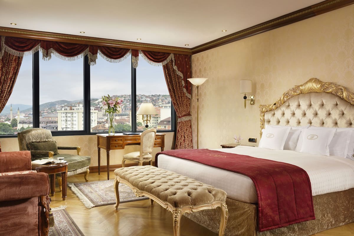 A luxurious hotel room with a large bed, a desk, a chair, and a view of the city through large windows.