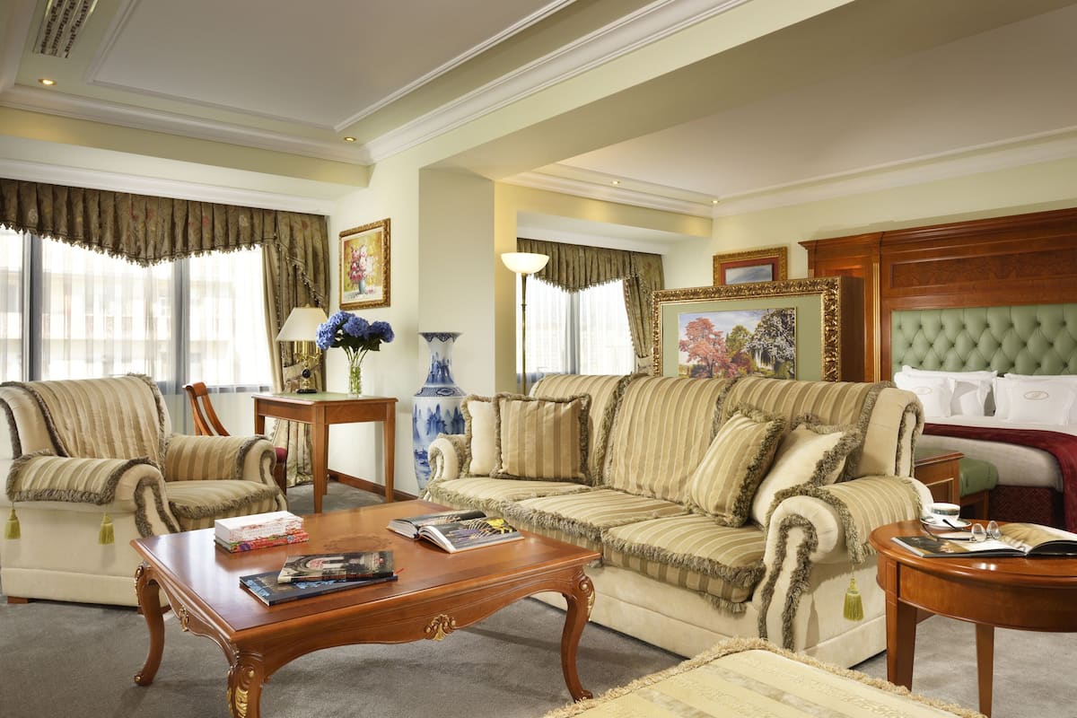 A spacious living room with a large sofa, a coffee table, and a reading area. There is a bed with a green headboard in the background.