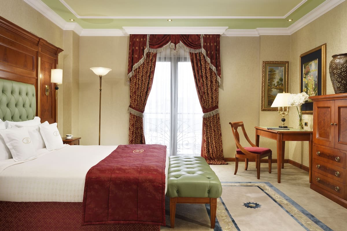 A hotel room with a large bed, a green upholstered bench, a wooden desk, and a chair. There is a window with curtains, a floor lamp, and a cabinet with drawers.