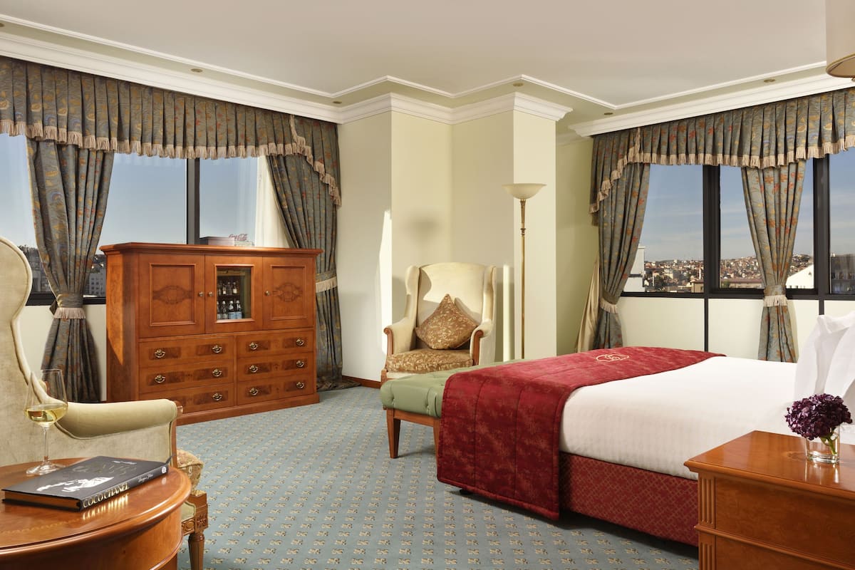 A hotel room with a large bed, a wooden dresser, a chair, a desk with a book and glass, and large windows with curtains.