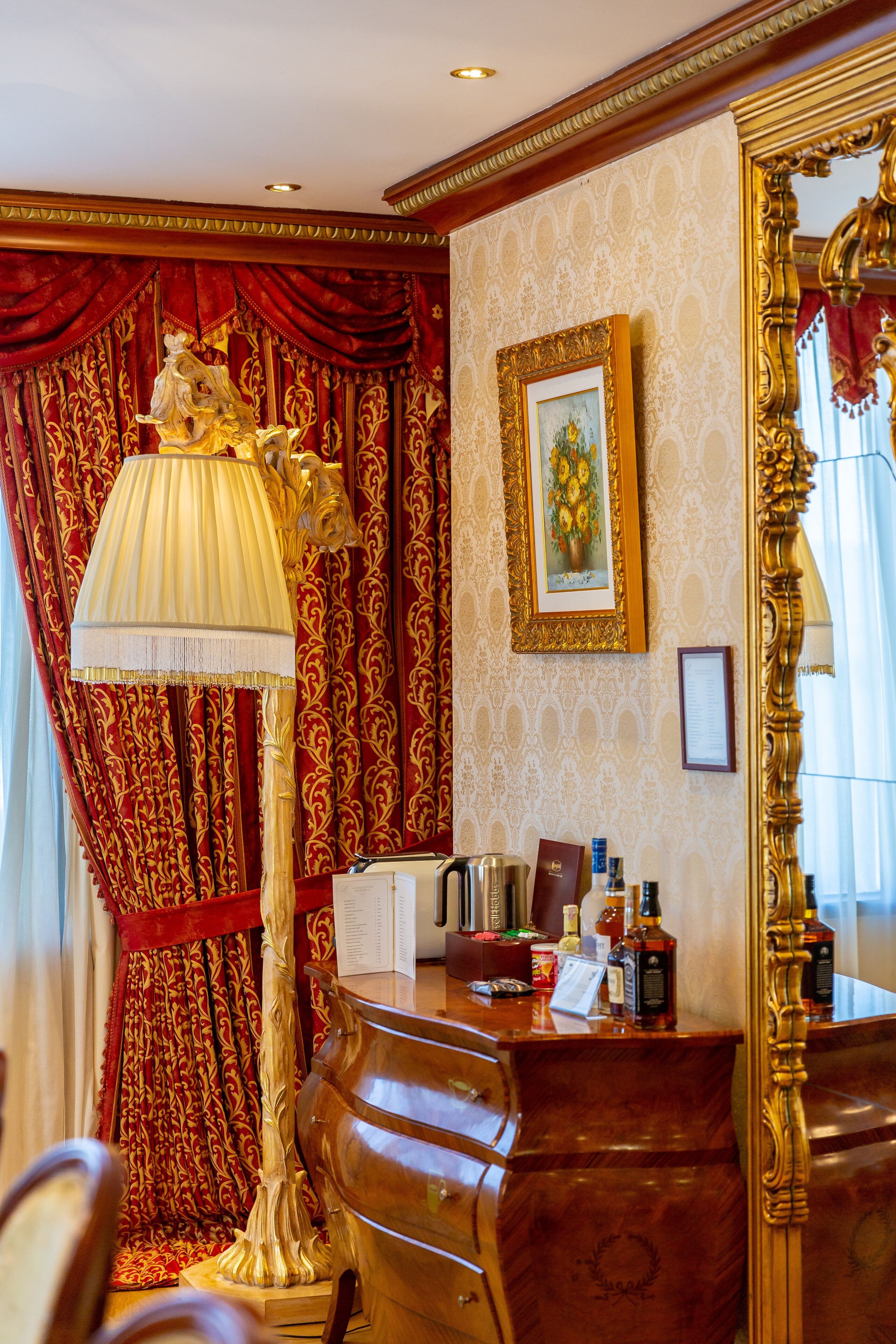 A room with a wooden desk, a large ornate lamp, red curtains, and a framed painting on the wall.