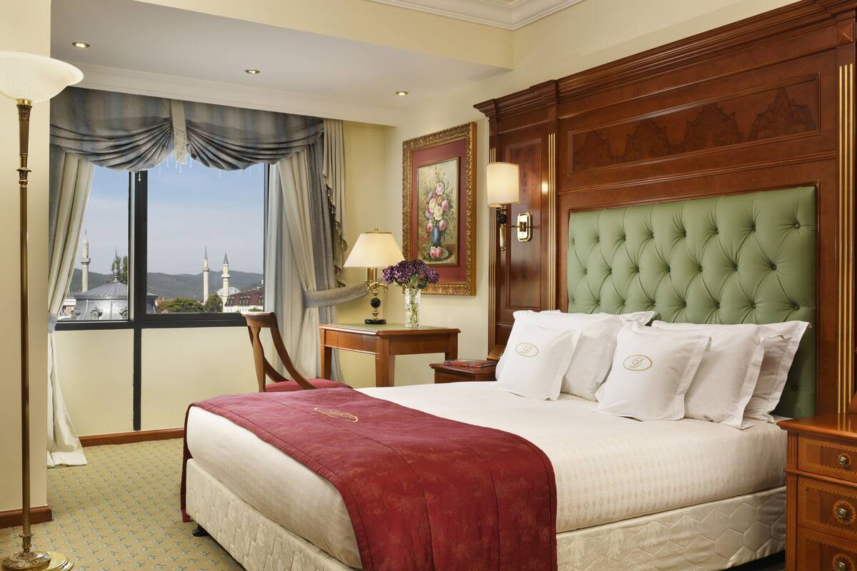 A hotel room with a large bed, a view of a cityscape, a wooden headboard, and a nightstand with a lamp.