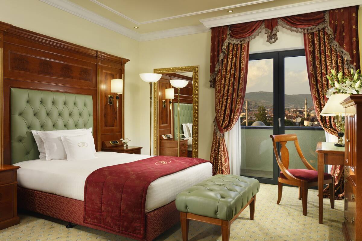 A hotel room with a large bed, a green upholstered bench, a wooden chair, a desk with a lamp, and a view of a cityscape through a large window.