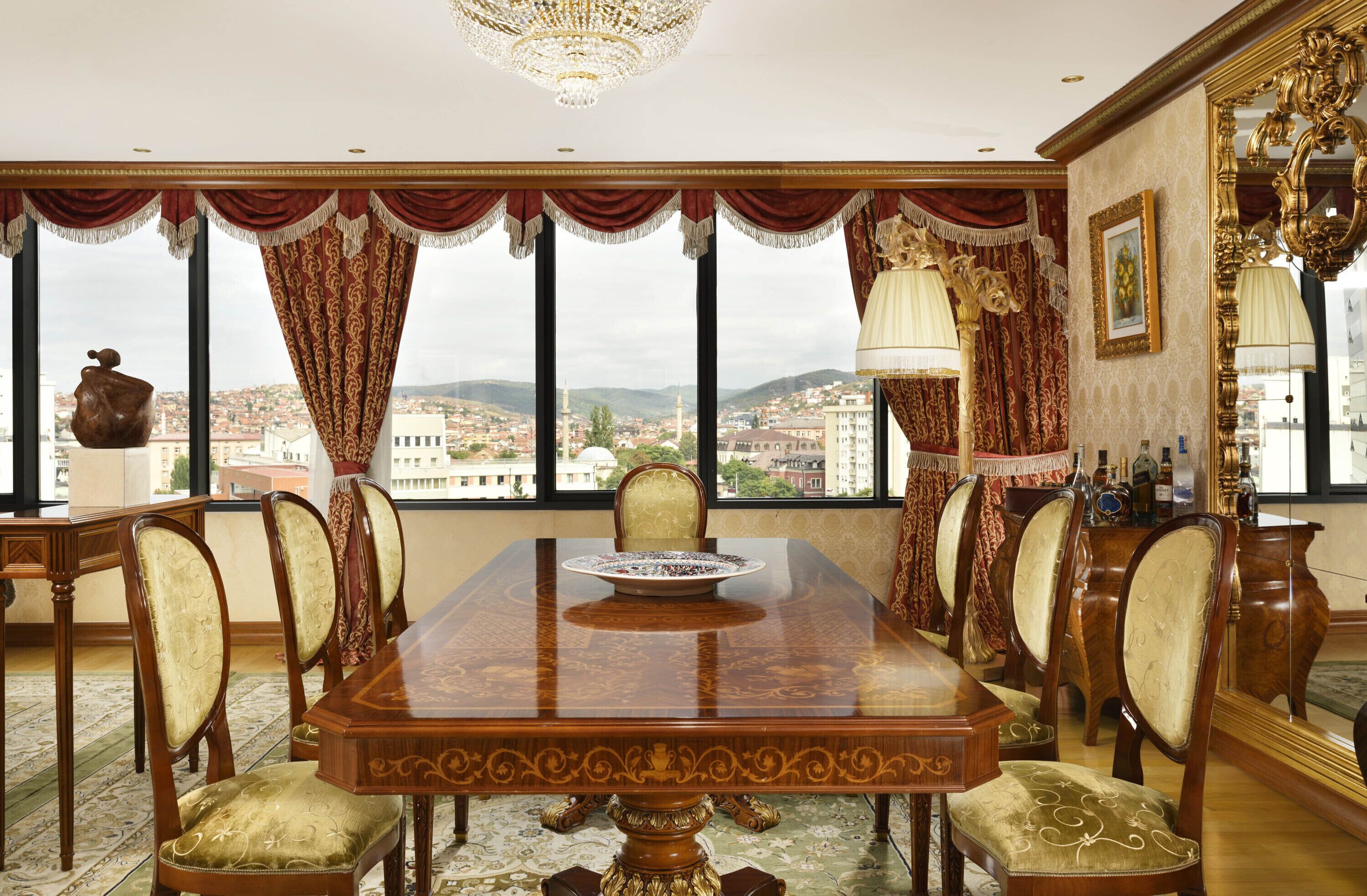 Ein Esszimmer mit einem großen Holztisch, verzierten Stühlen und Blick auf eine Stadtlandschaft durch große Fenster.