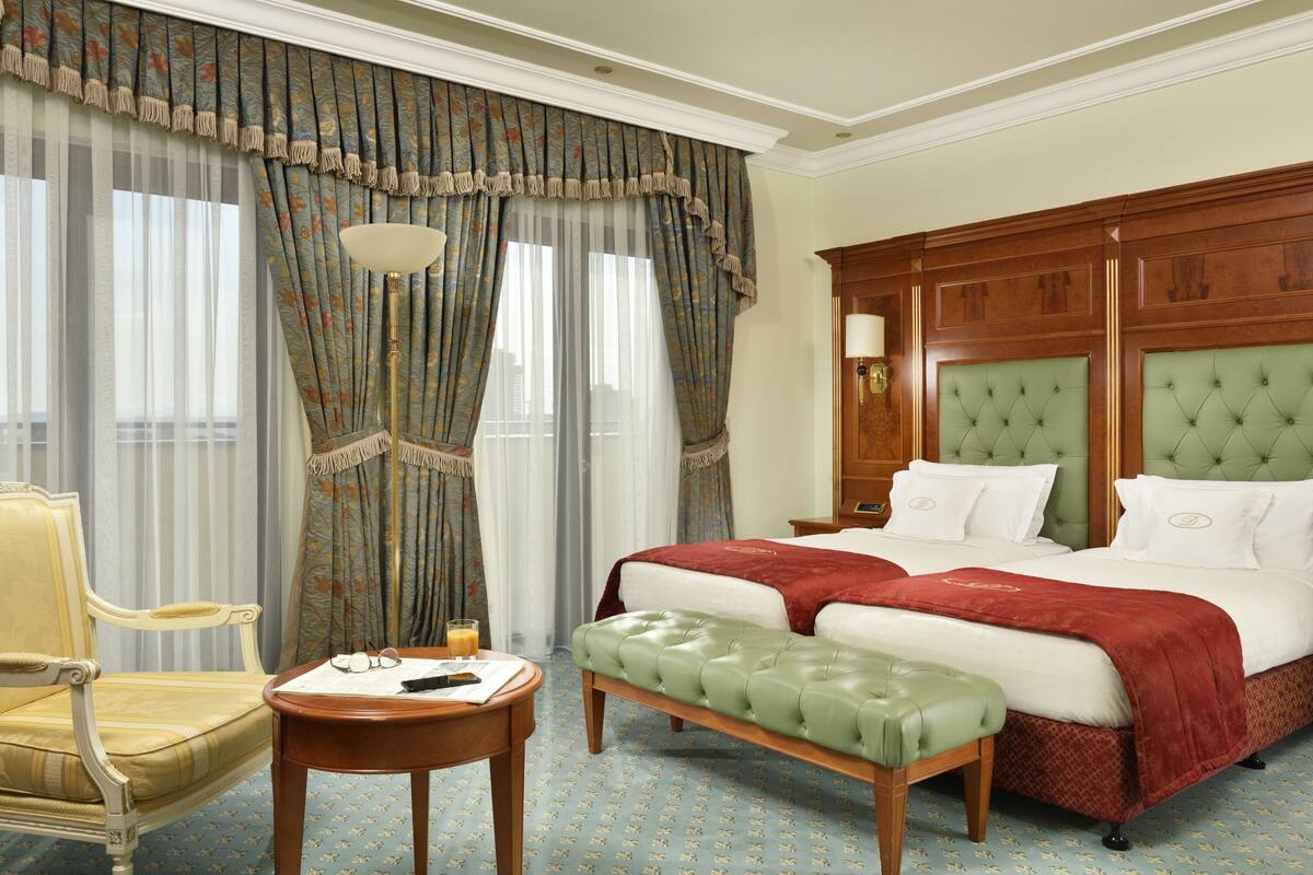 A hotel room with a large bed, a green upholstered bench, a wooden nightstand, a chair, and a table with a lamp.