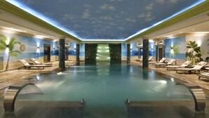 A large indoor swimming pool with lounge chairs and palm trees, surrounded by walls with a sky and clouds mural. - Swiss Diamond Hotel Prishtina (Pristina)