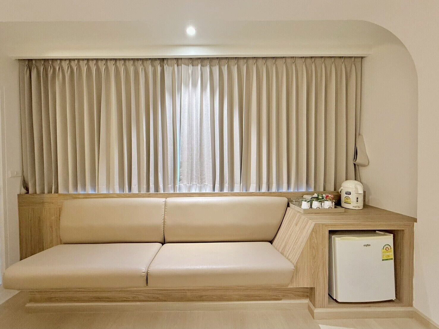 Une pièce avec un canapé beige, une table d’appoint en bois, un réfrigérateur blanc et une fenêtre ornée de rideaux beiges.