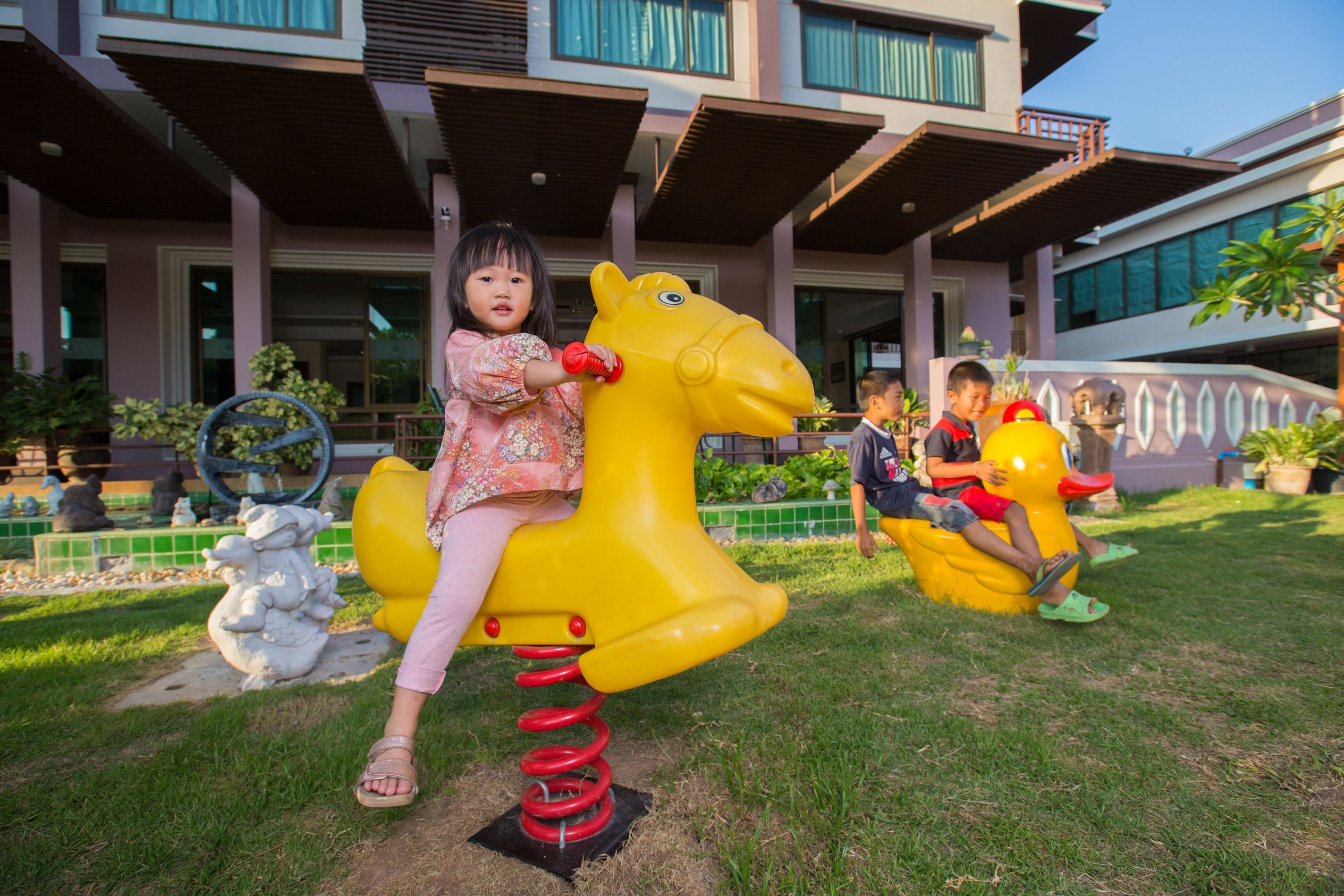 A child on a yellow horse-shaped swing set in a grassy area with a building in the background.