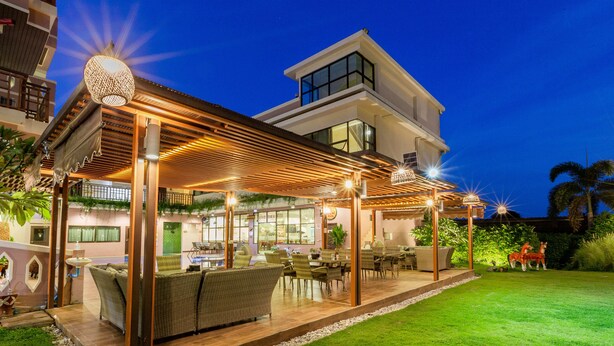 Un comedor al aire libre moderno con mobiliario de mimbre y una pérgola de madera, rodeado de un césped cuidado y palmeras.