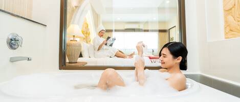 A woman relaxing in a bathtub with a view of a living room where a person is reading a book.