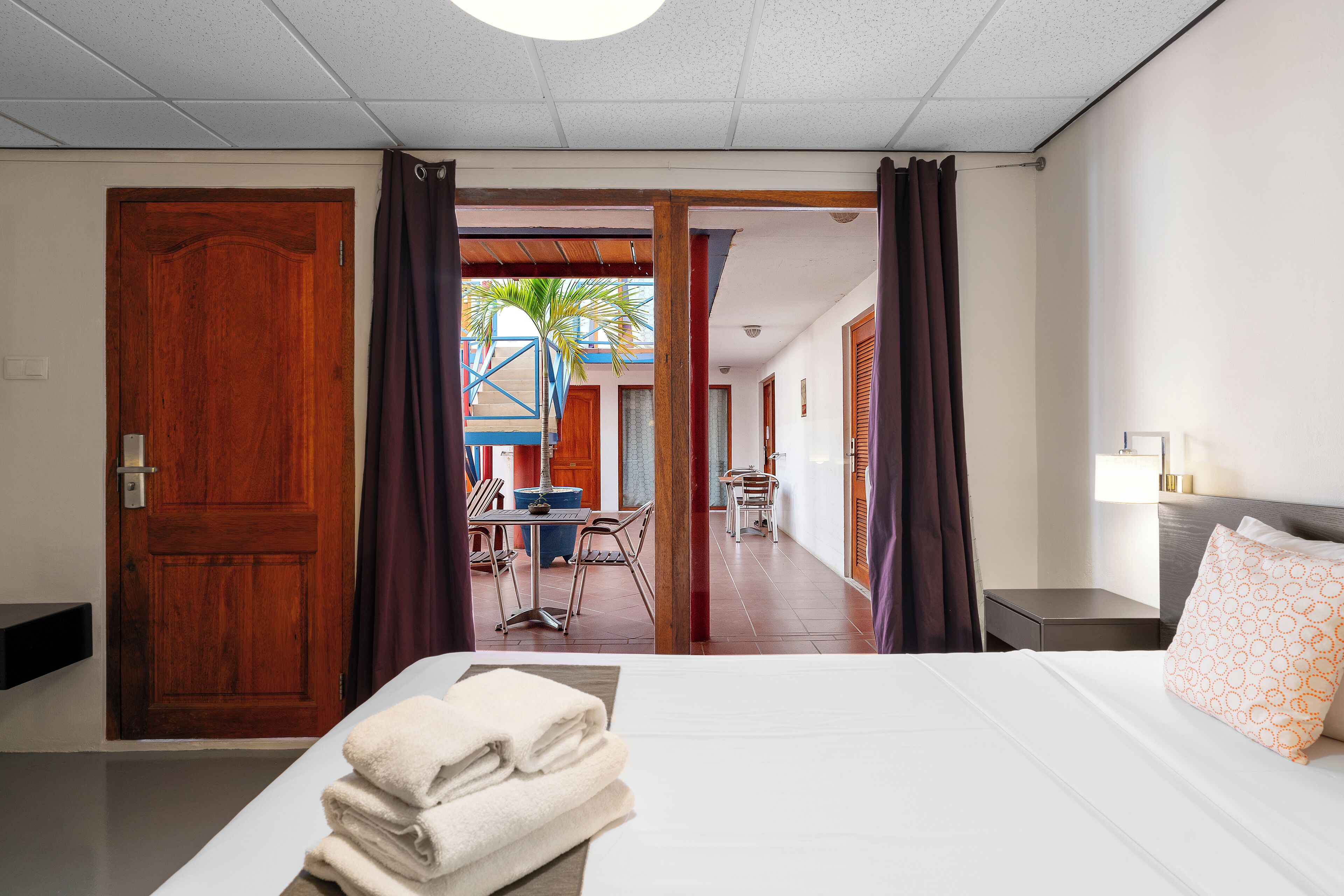 Habitación de hotel con cama, mesita de noche, lámpara y vistas al exterior por una puerta abierta.