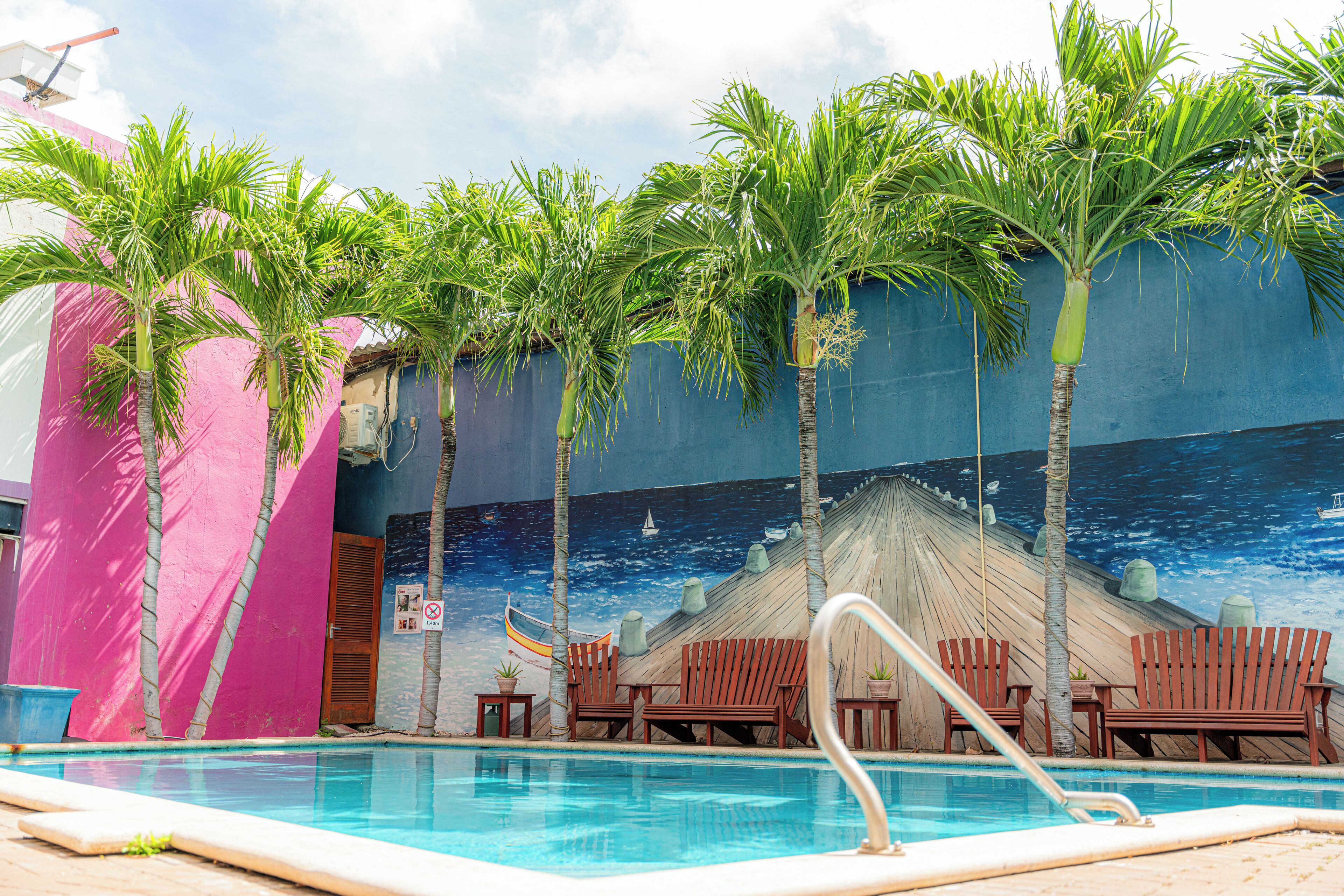 A pool area with palm trees, a mural, and wooden chairs.