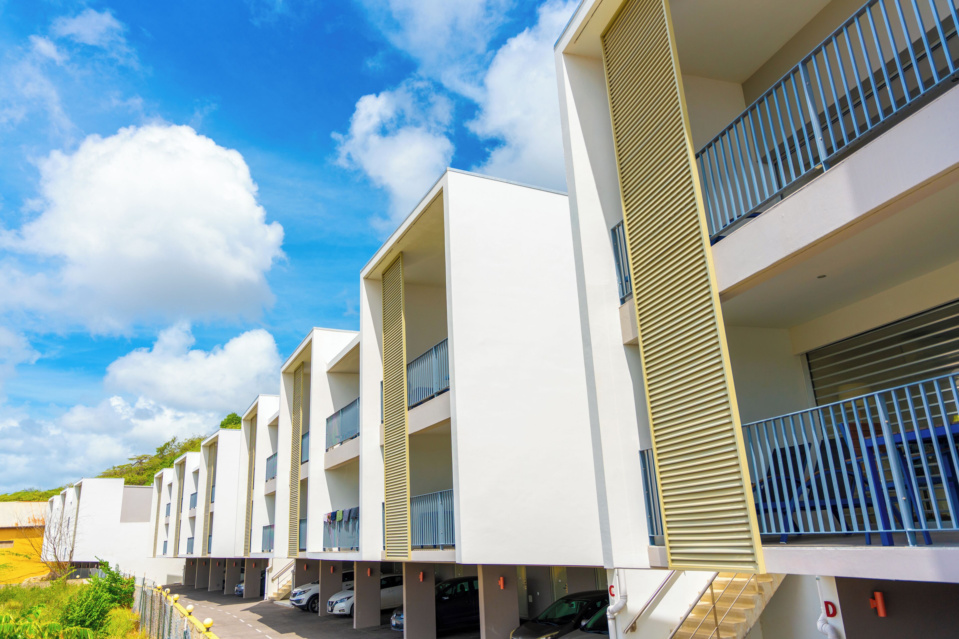 A modern multi-story building with balconies, a parking area, and a clear sky.