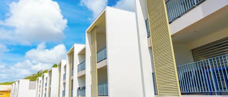 A modern multi-story building with balconies, a parking area, and a clear sky.