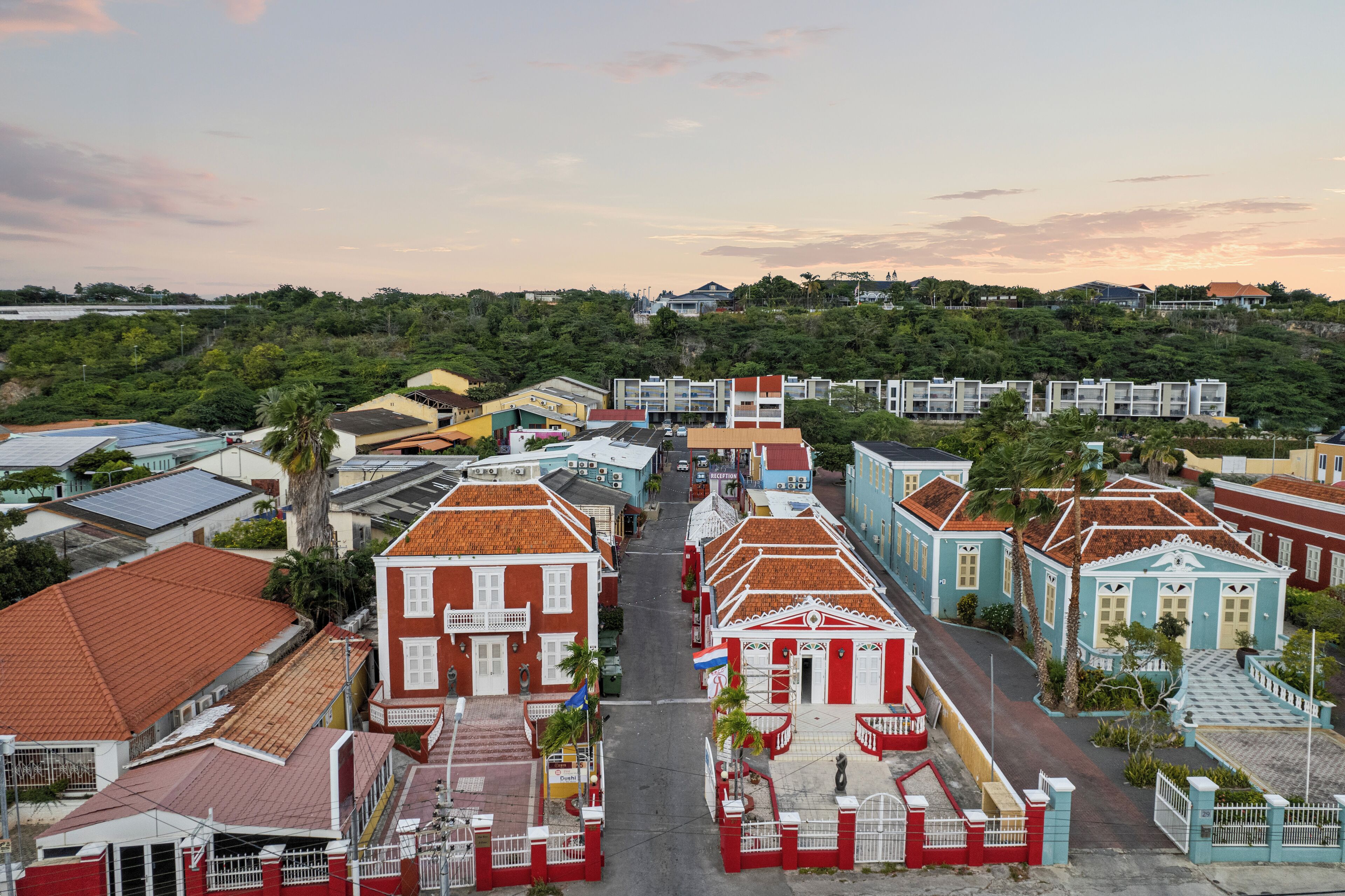 A coastal town with buildings featuring red and white facades, a central street, and a backdrop of greenery and hills.