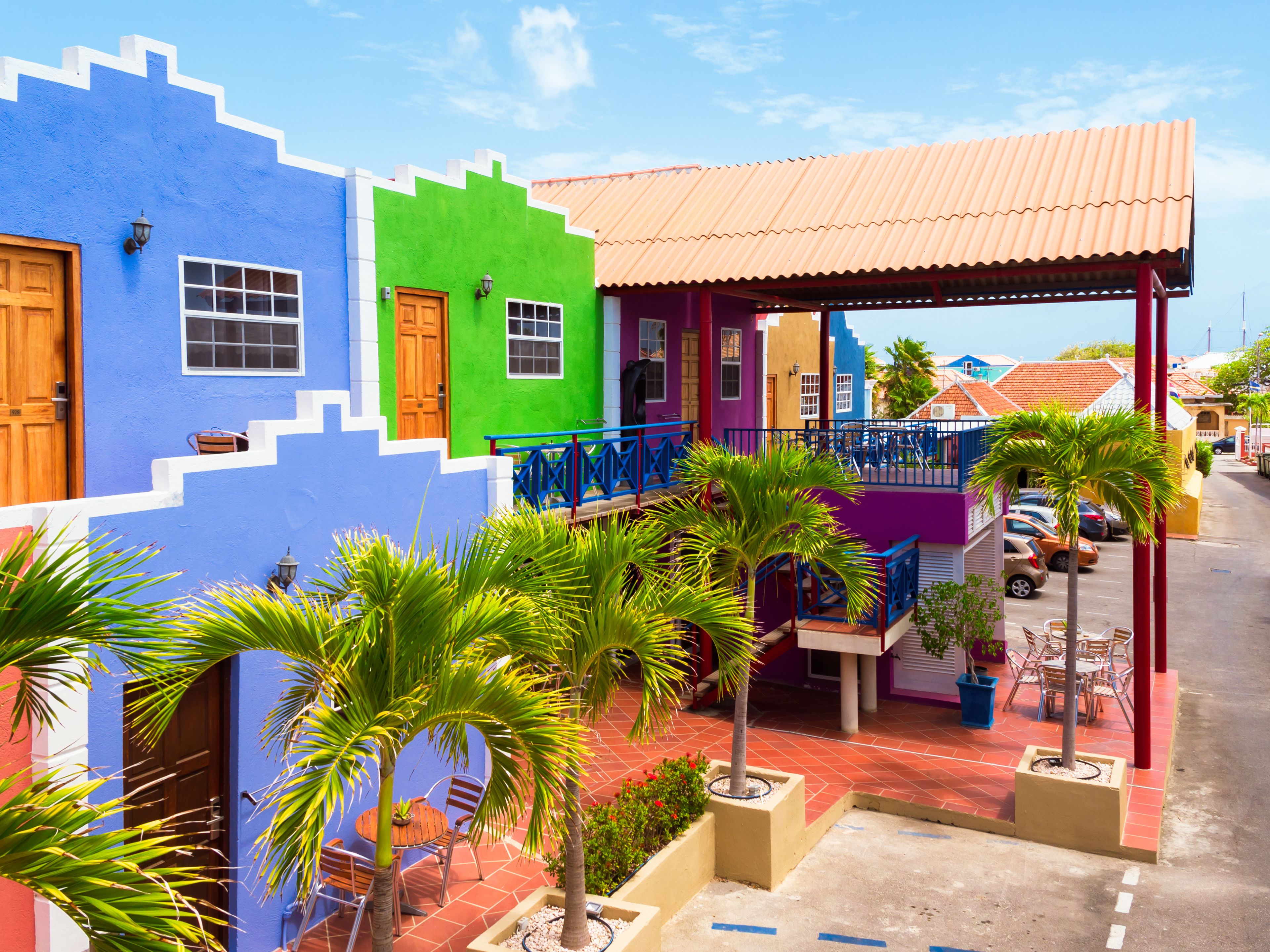 A colorful building with a blue, green, and purple facade, a red roof, and a small outdoor seating area with chairs and tables.