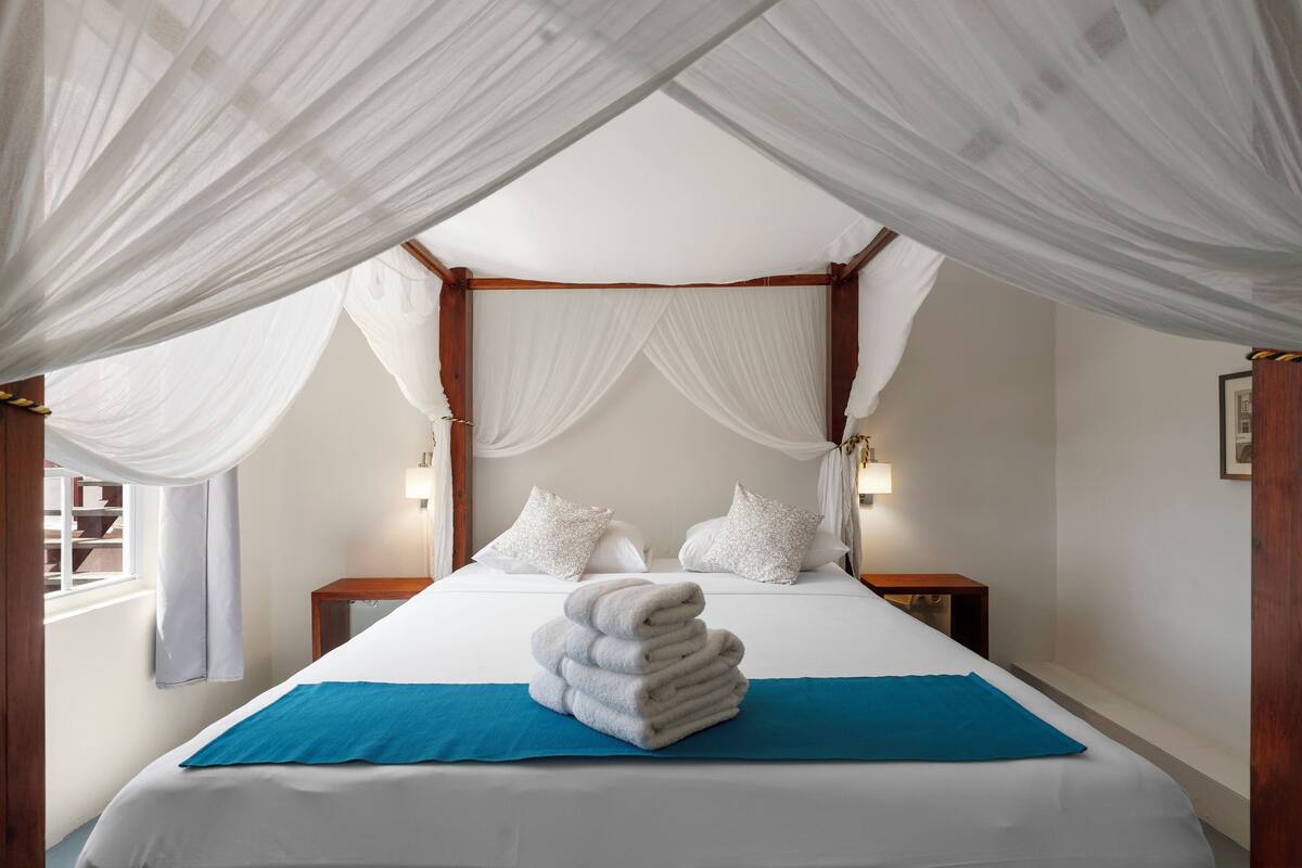 Una cama con dosel y cuatro columnas con ropa blanca y una pila de toallas sobre un corredor azul para la cama.