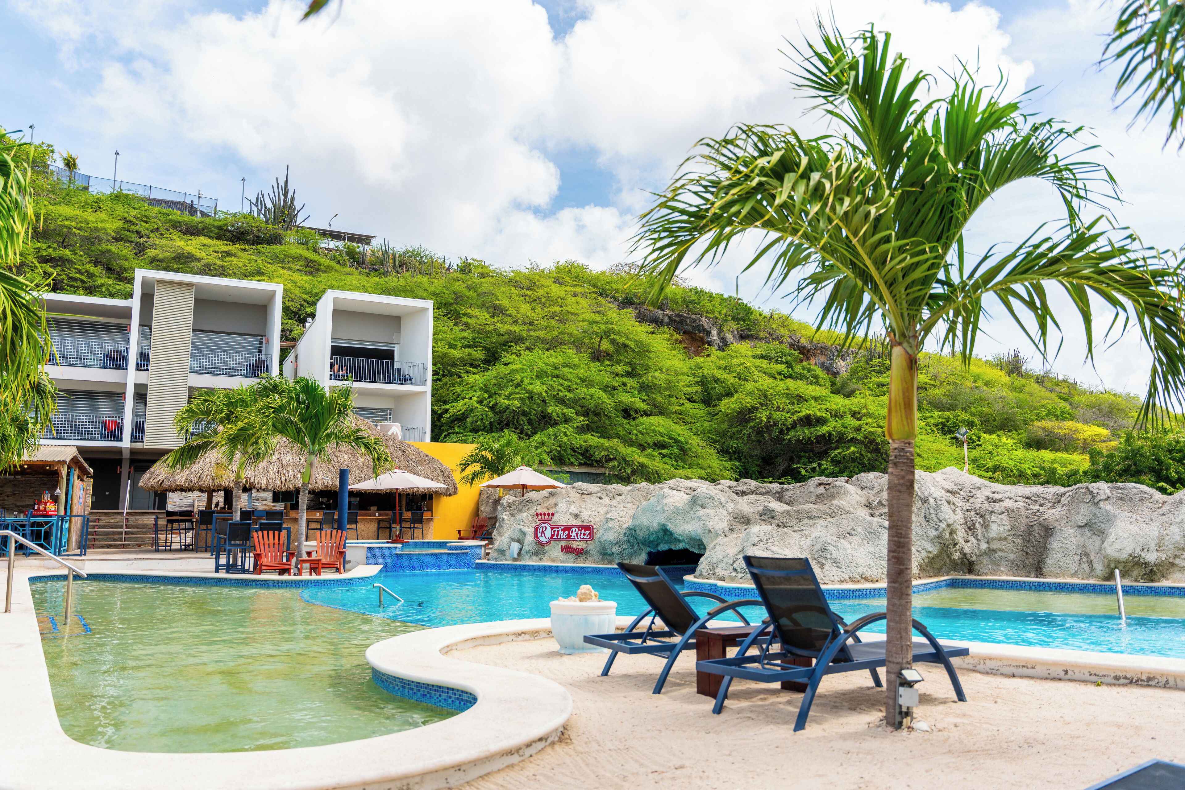 Un espace piscine avec des chaises longues, un palmier et un bâtiment sur lequel on peut lire l’inscription « The Ritz Village ».