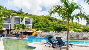 Un espace piscine avec des chaises longues, un palmier et un bâtiment sur lequel on peut lire l’inscription « The Ritz Village ».