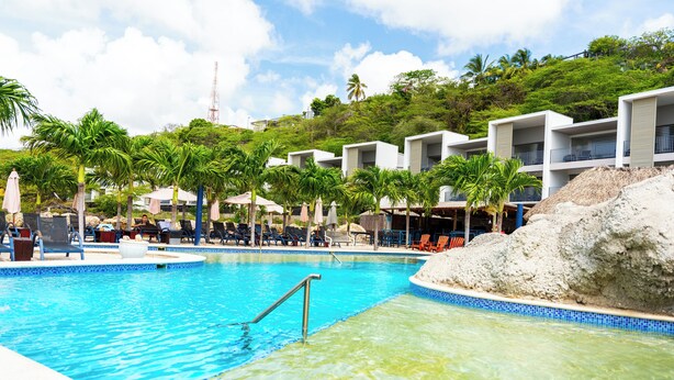 A swimming pool with clear water, surrounded by lounge chairs and umbrellas, with modern buildings and palm trees in the background. - The Ritz Village Hotel  (Willemstad)