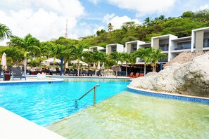 Una piscina con agua cristalina, rodeada de tumbonas y sombrillas, con edificios modernos y palmeras al fondo.