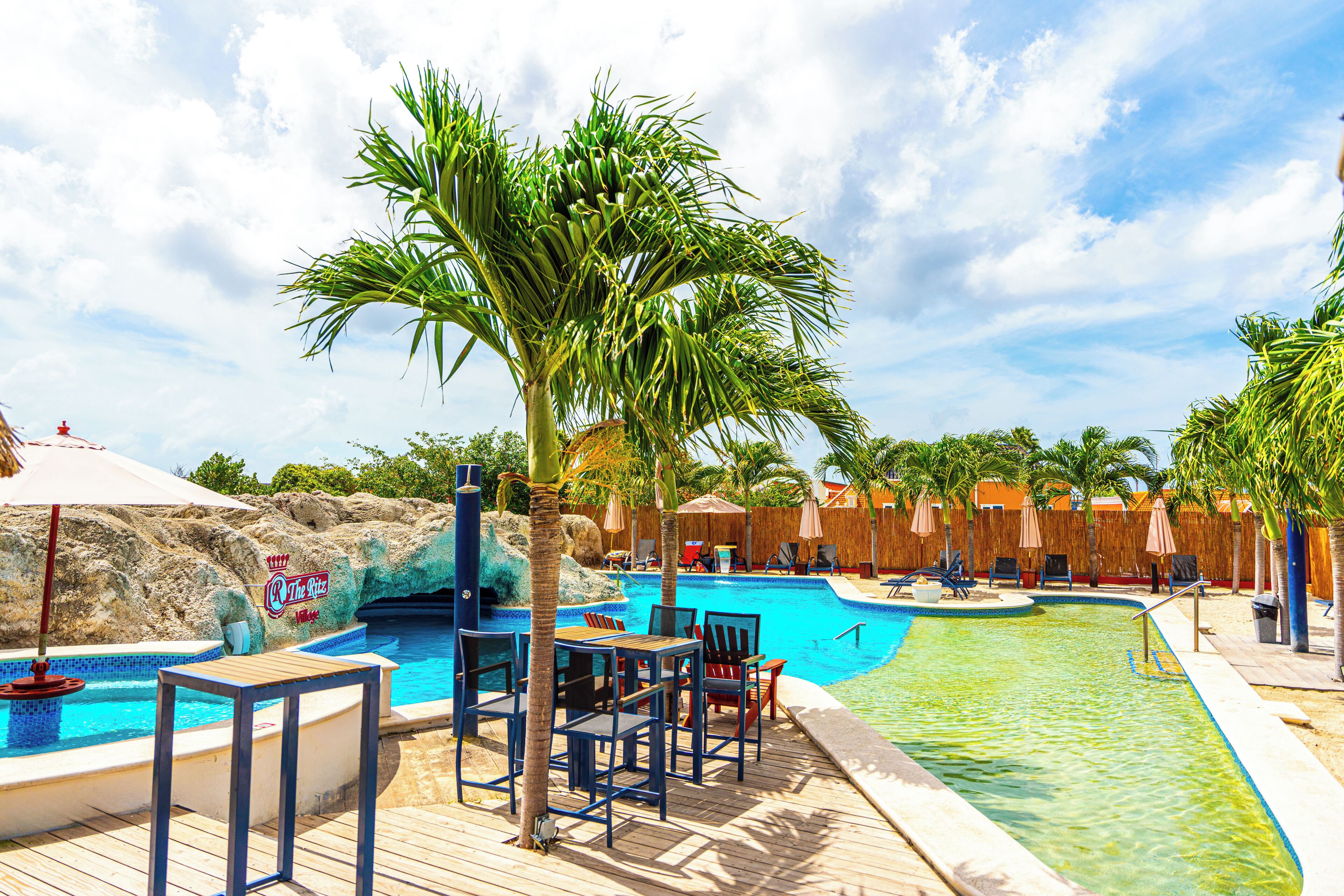 A swimming pool area with lounge chairs and tables, surrounded by palm trees and a wooden deck.