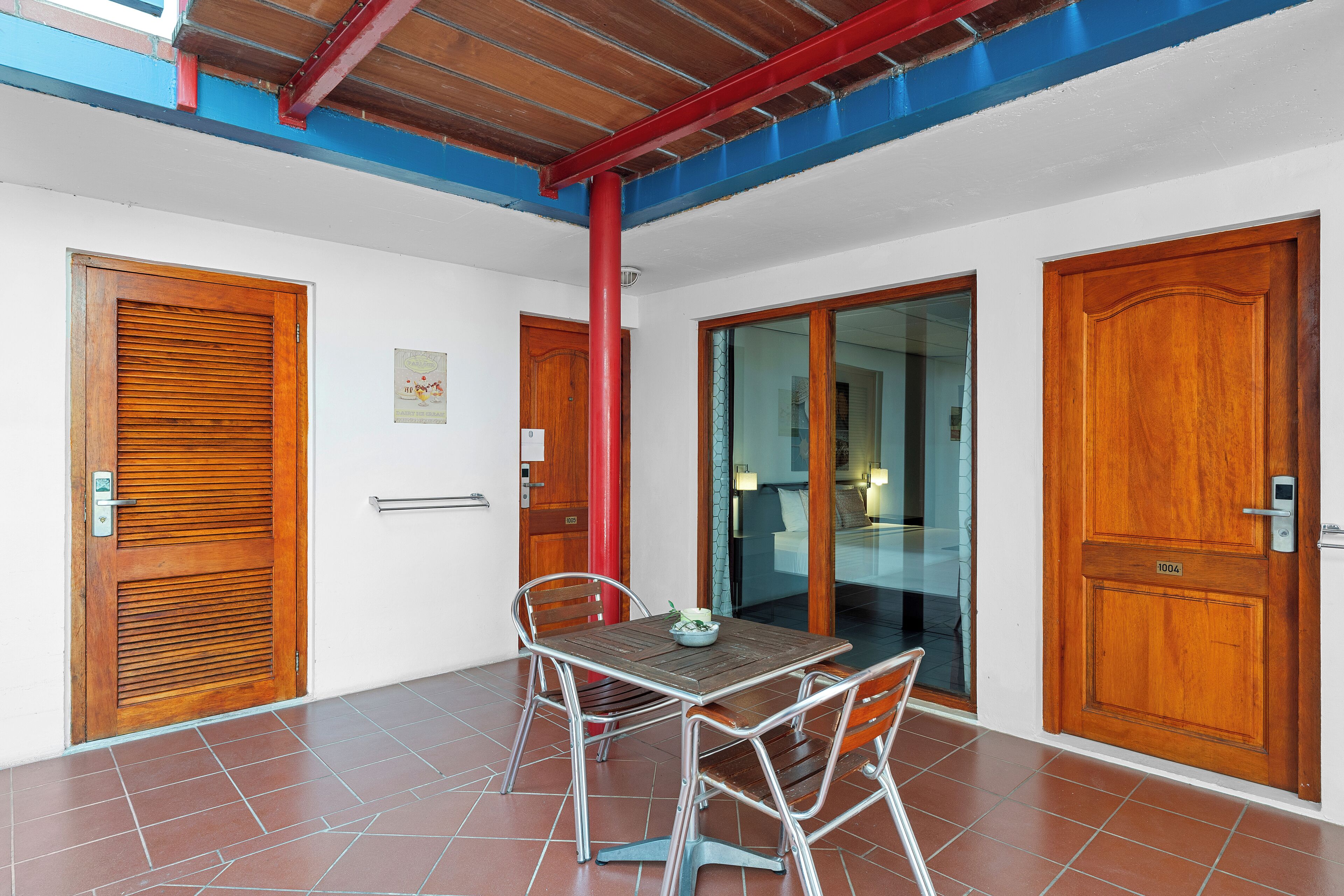 Una habitación con puerta de madera, puerta de cristal, una mesa de metal y sillas, y un pilar rojo.