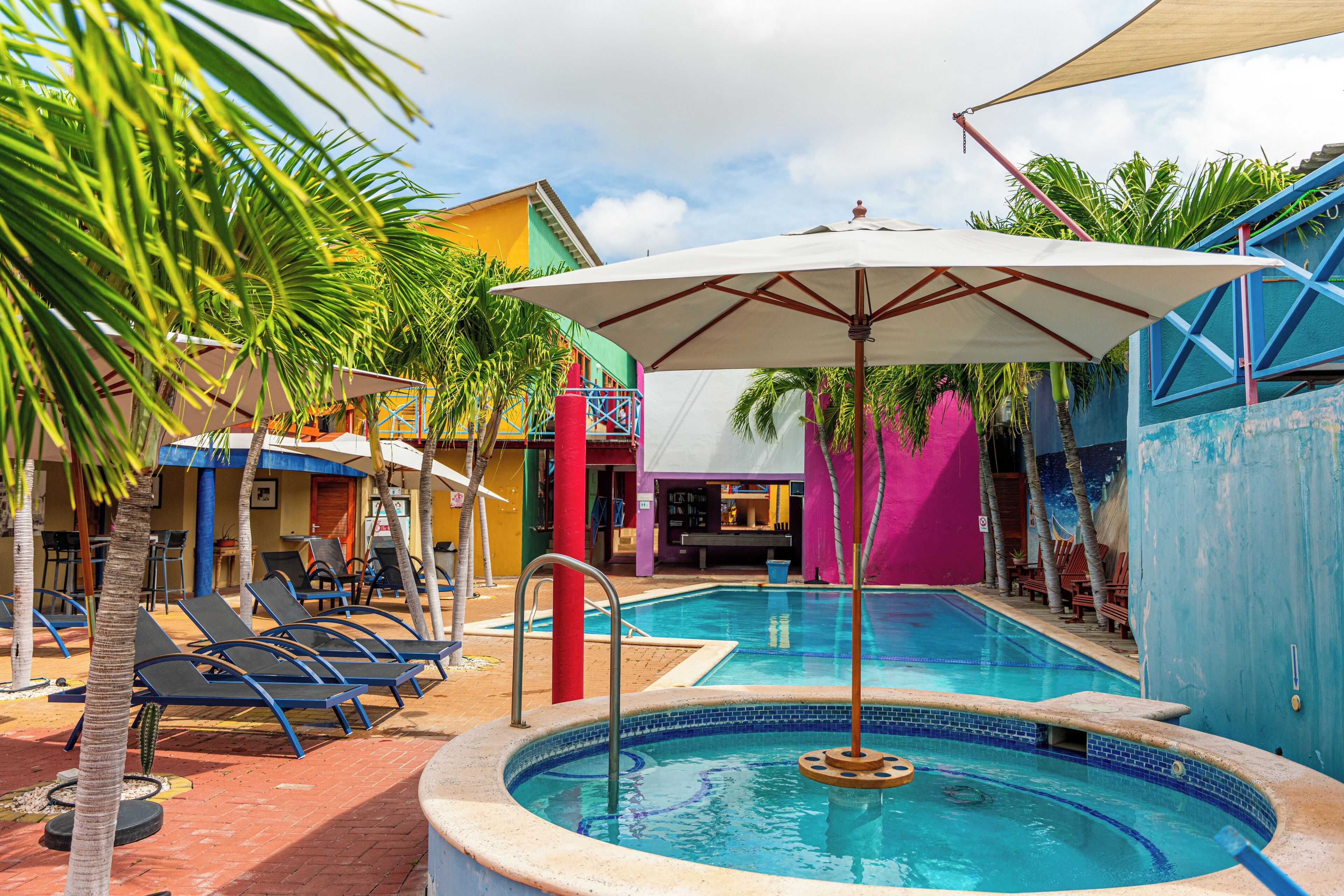 A swimming pool with lounge chairs and umbrellas, surrounded by colorful buildings and palm trees.