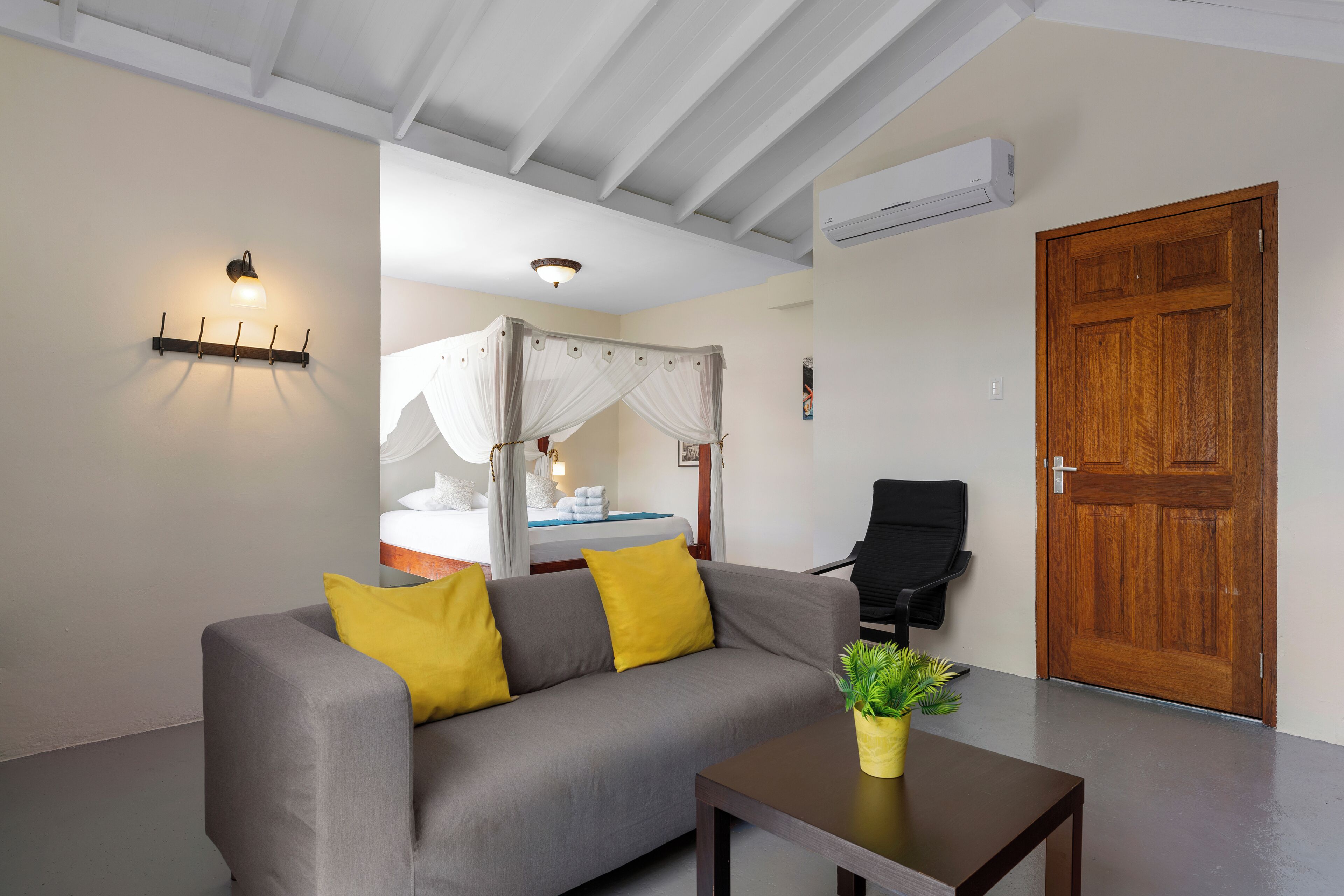 A modern room with a grey sofa, yellow cushions, a wooden table with a plant, a wooden door, and a bed with a canopy.