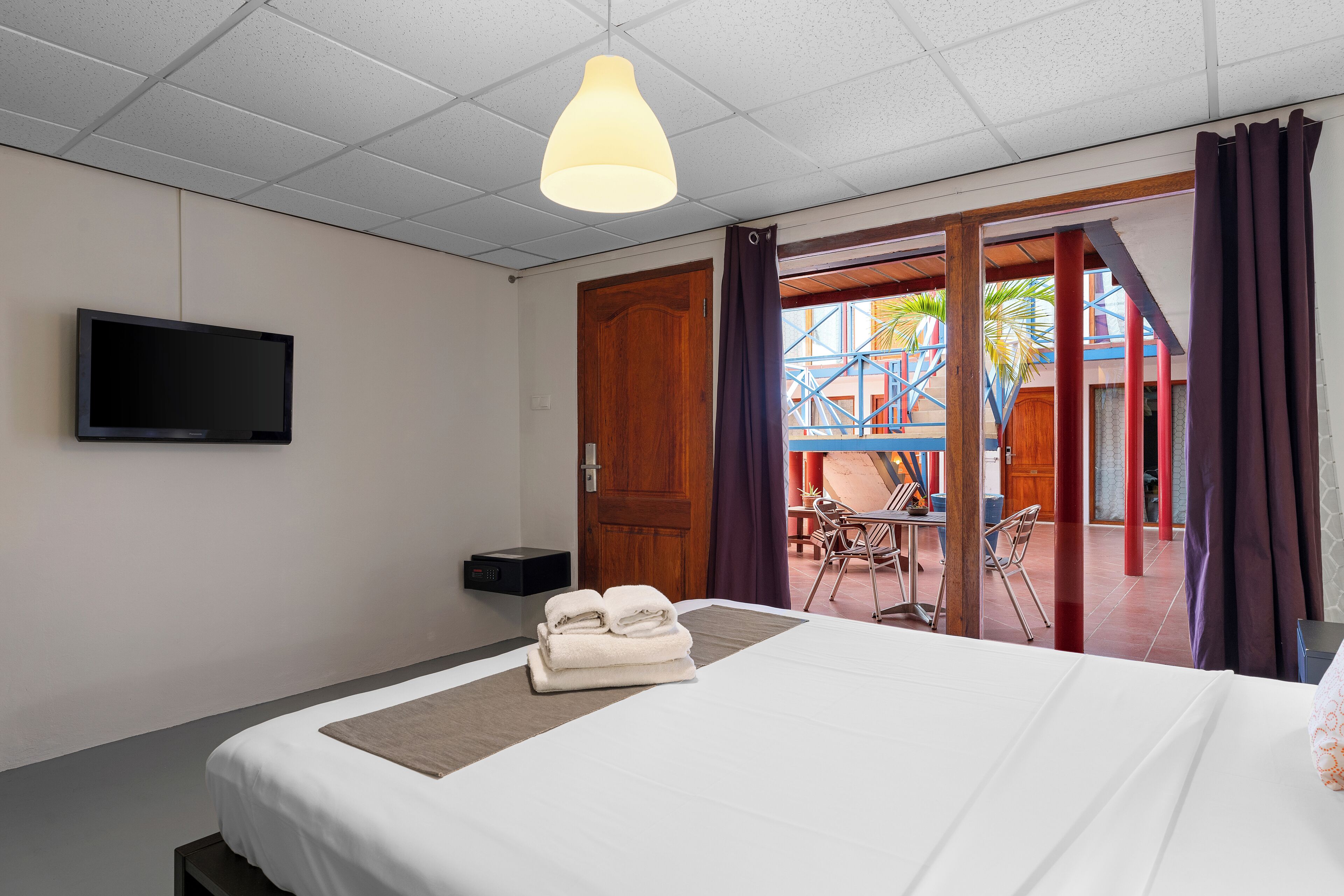 A hotel room with a bed, a flat-screen TV, a desk with a chair, and a balcony with red pillars and a view of the outside.