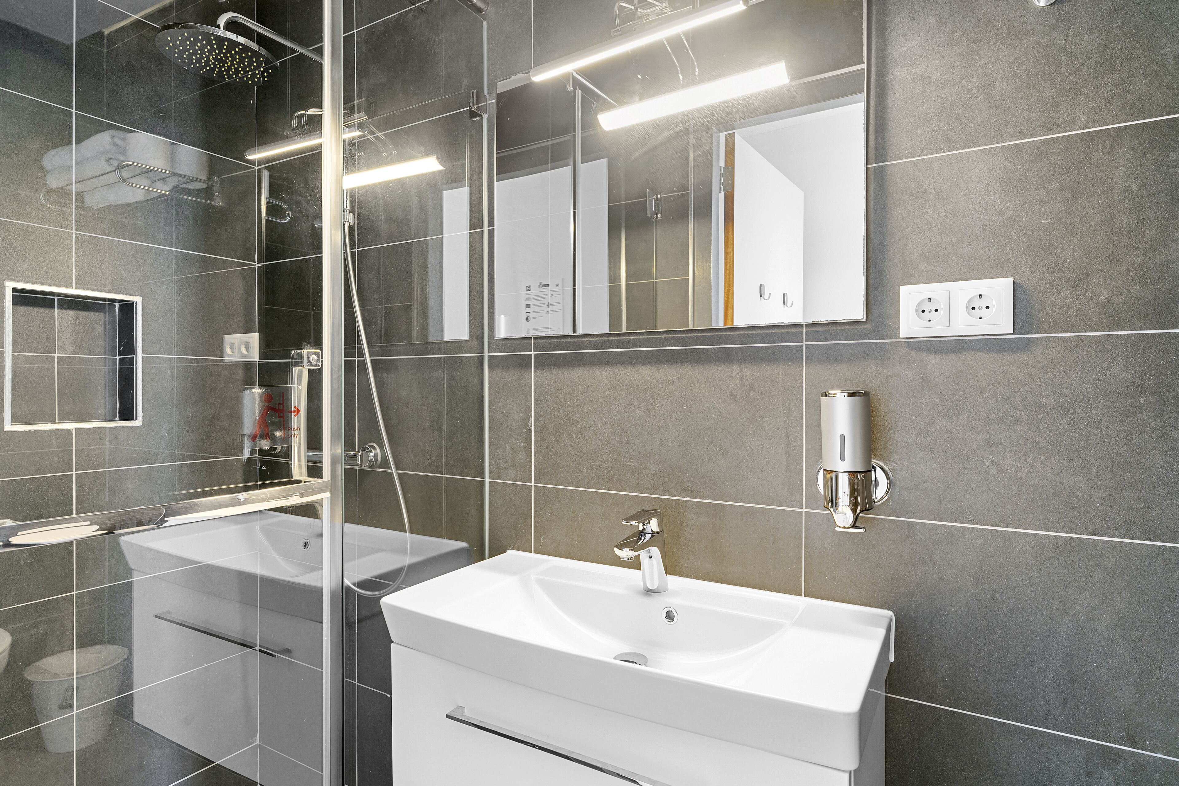 A modern bathroom with a glass-enclosed shower, two sinks, a large mirror, and a wall-mounted soap dispenser.