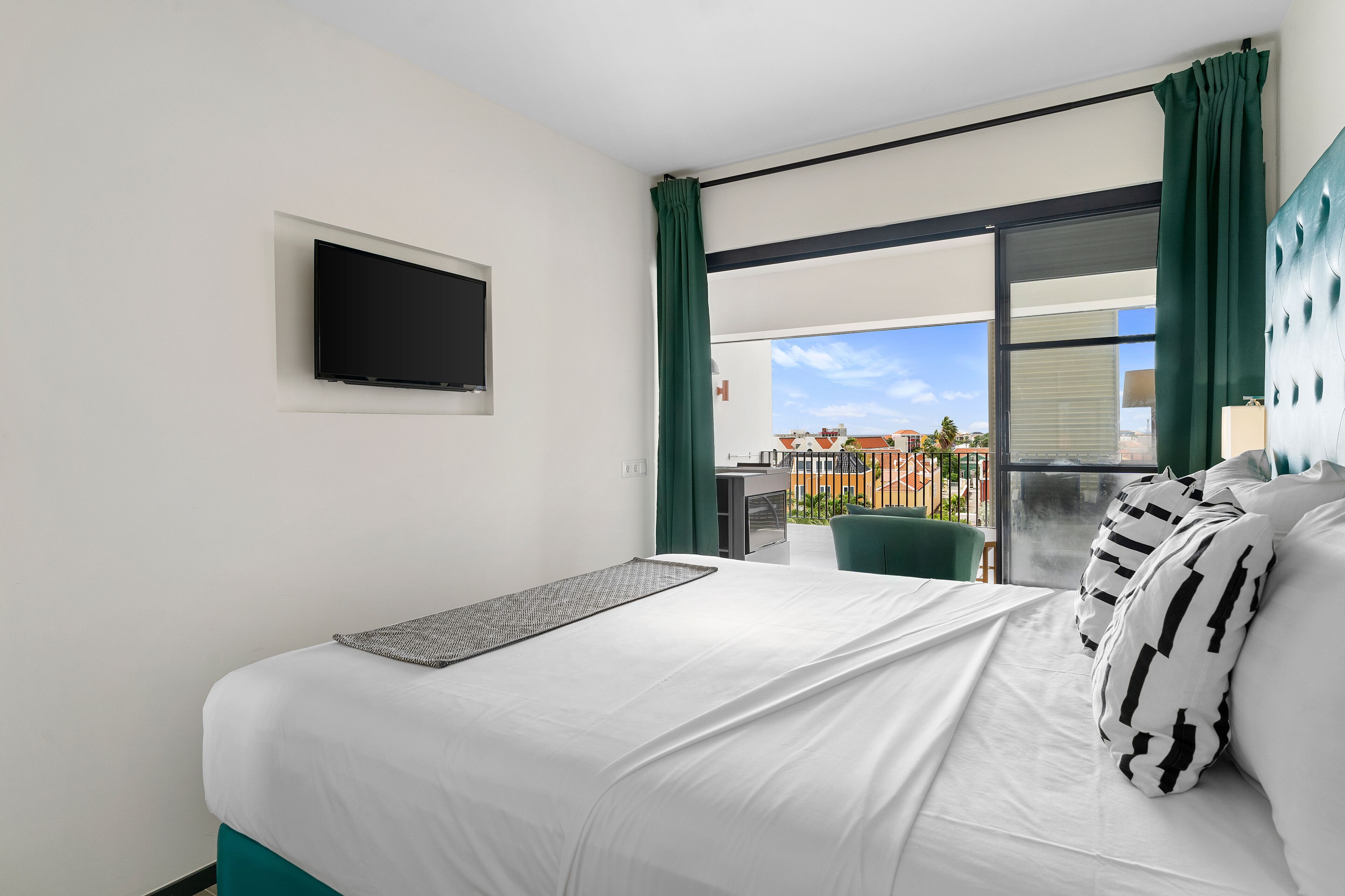 A modern hotel room with a bed, a flat-screen TV mounted on the wall, and a balcony with a view of the city.