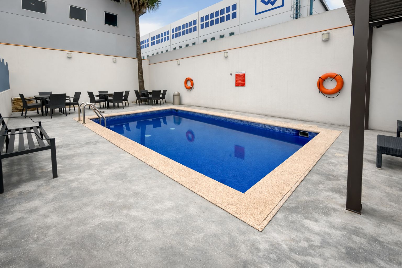 A swimming pool with a tiled edge, surrounded by a concrete deck and black chairs.