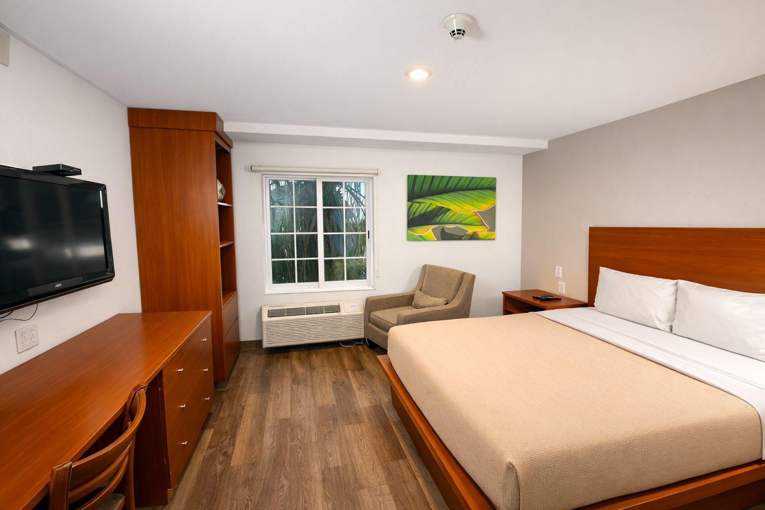 A hotel room with a bed, a television, a chair, a window, and a wall-mounted artwork.