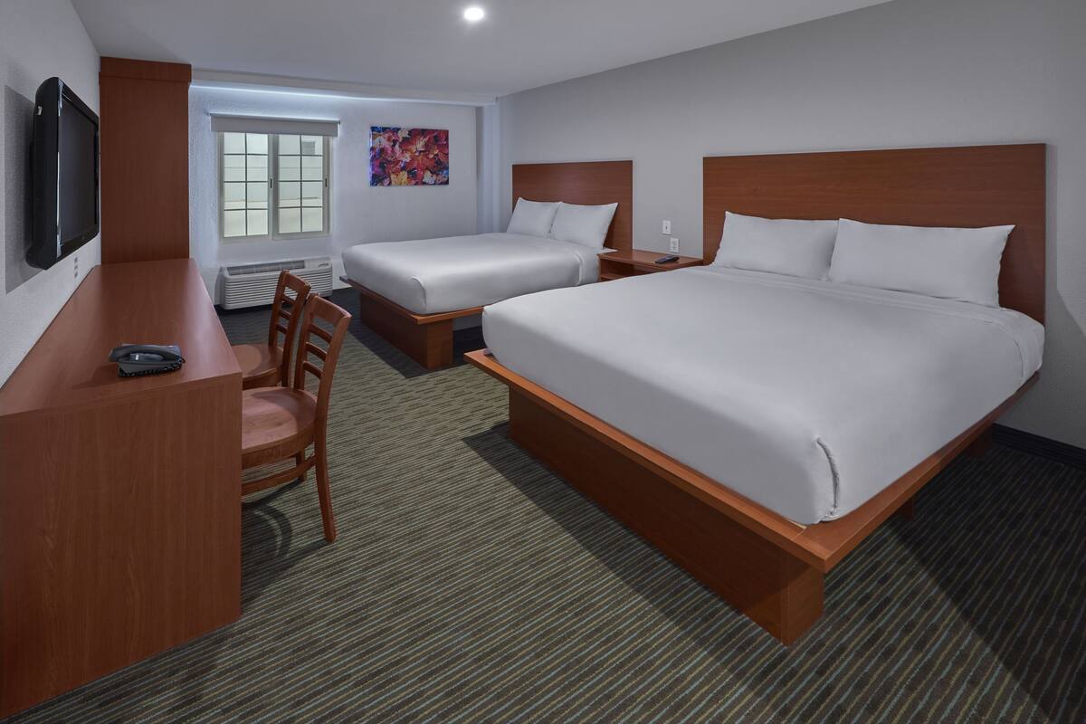 A hotel room with two single beds, a larger double bed, a desk with a chair, a television, and a window with blinds.
