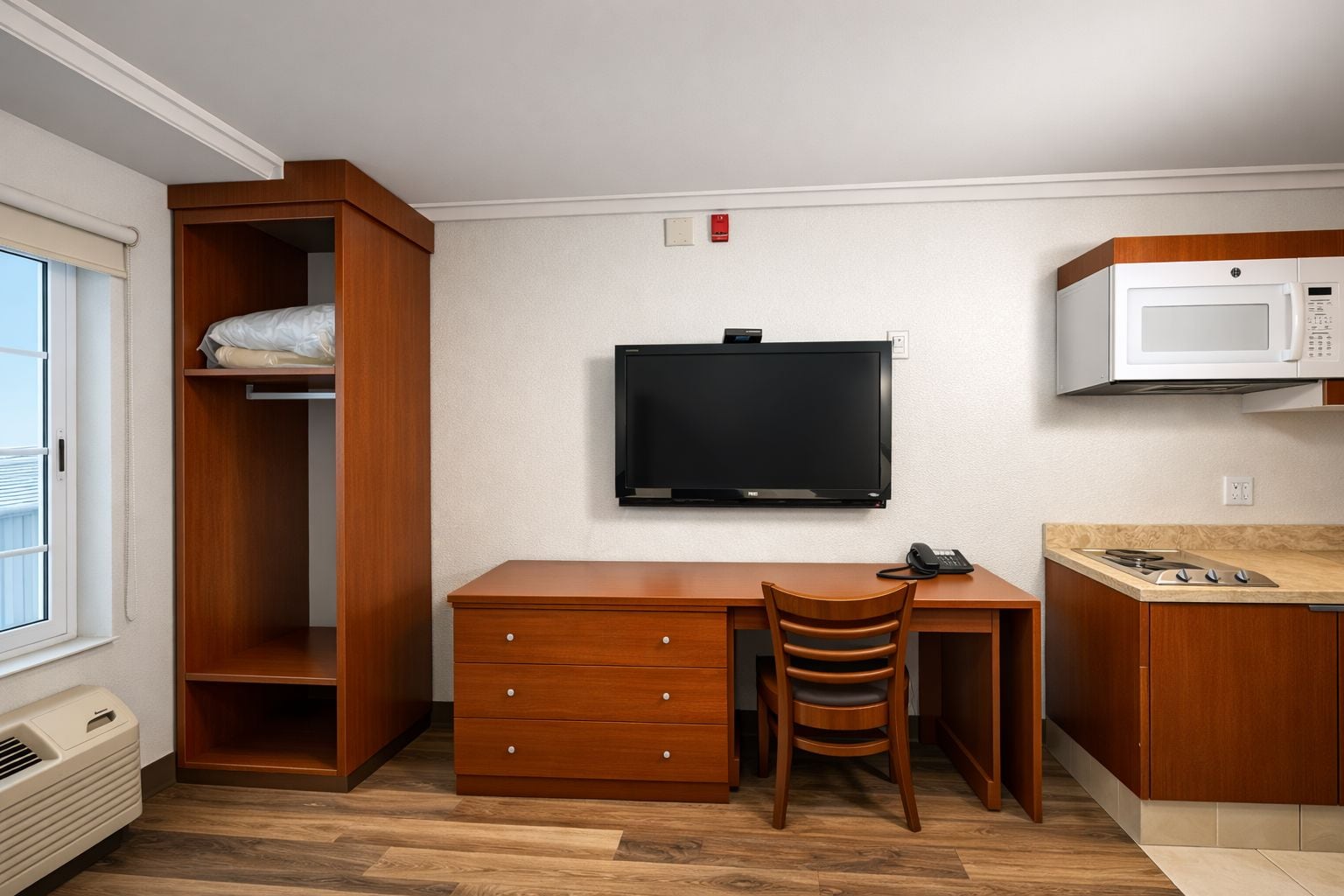 A compact room with a wooden desk, a television, a microwave, and a small kitchenette.