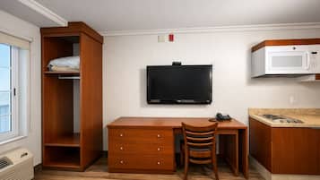 A compact room with a wooden desk, a television, a microwave, and a small kitchenette.